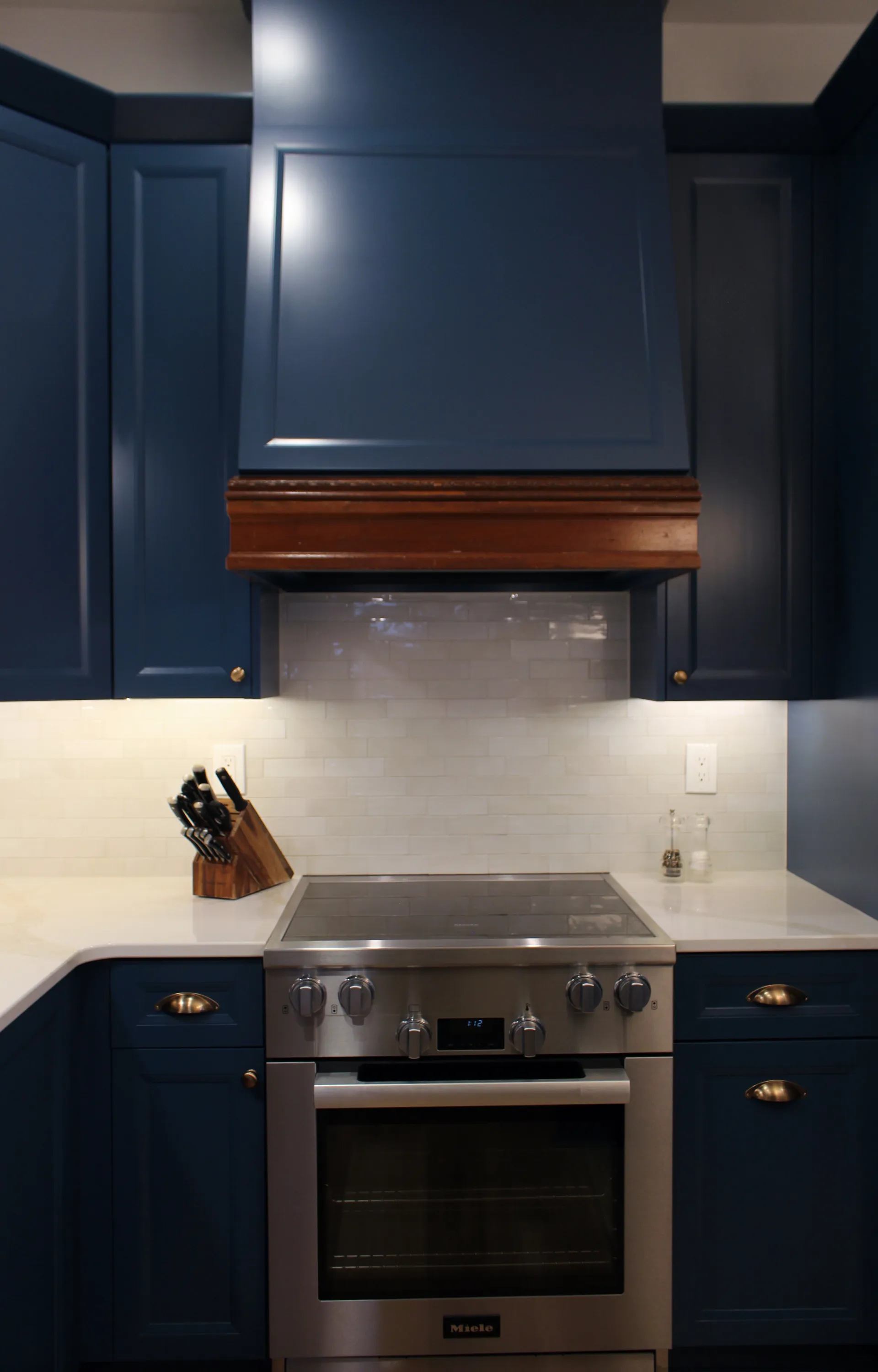 Sleek kitchen with deep blue cabinets, stainless steel oven, and wooden accents on the range hood. A knife block and bright lighting add a modern touch.
