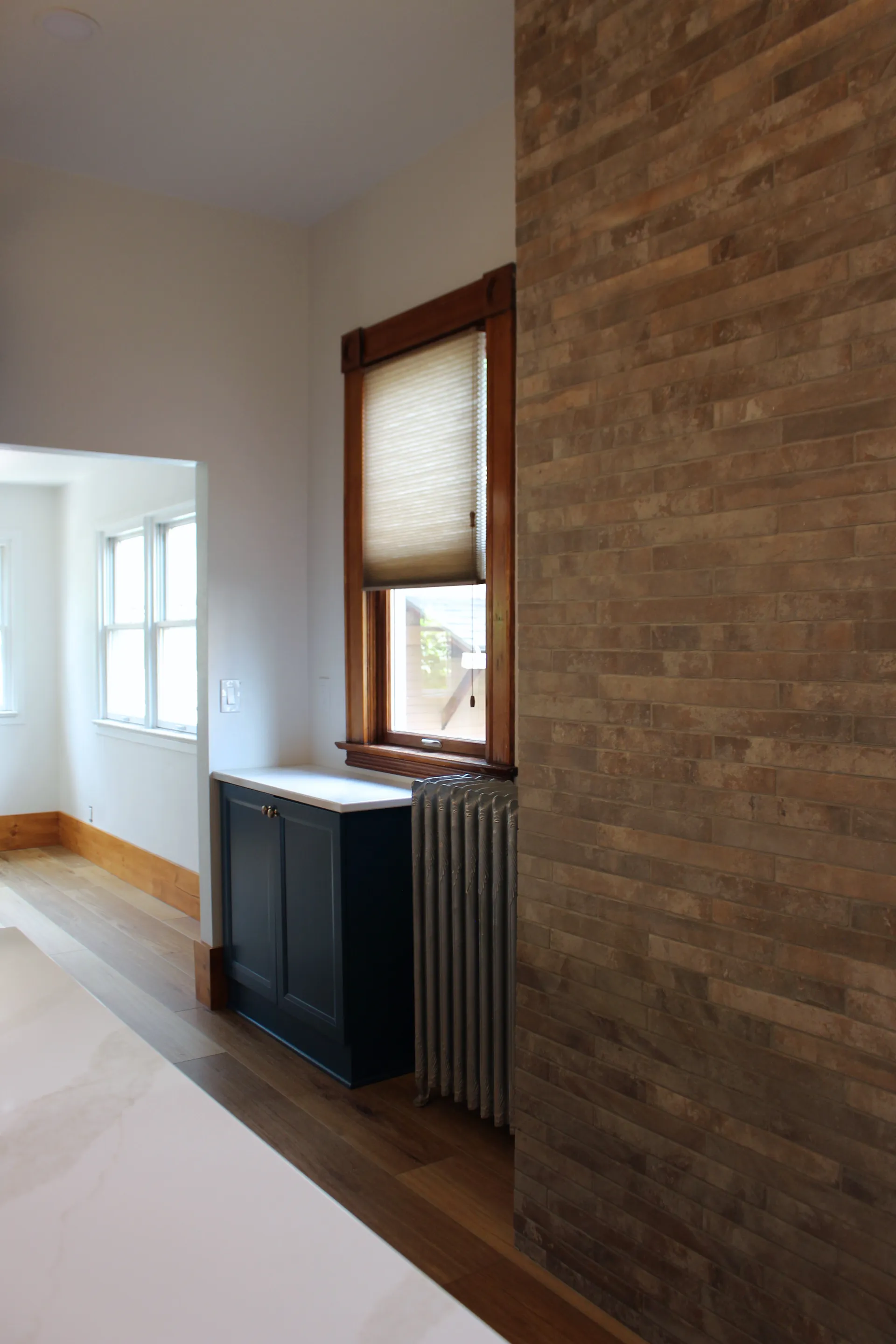 A modern interior features a tall brick wall on the right, a wooden floor, and a small blue cabinet under a window with blinds. Light streams from another room, creating a warm, cozy atmosphere.