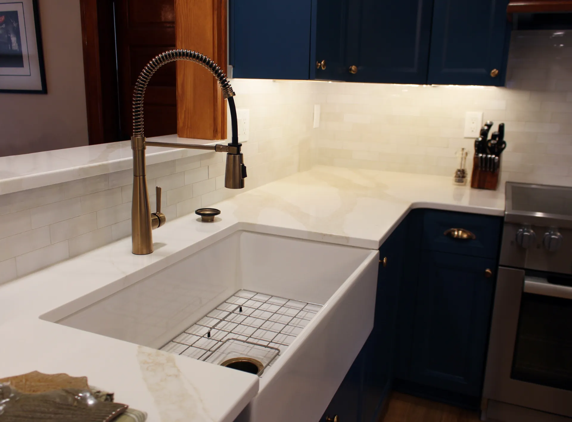 Modern kitchen with a white farmhouse sink, bronze faucet, and elegant white countertop. Blue cabinets add contrast, alongside a knife block and stove.