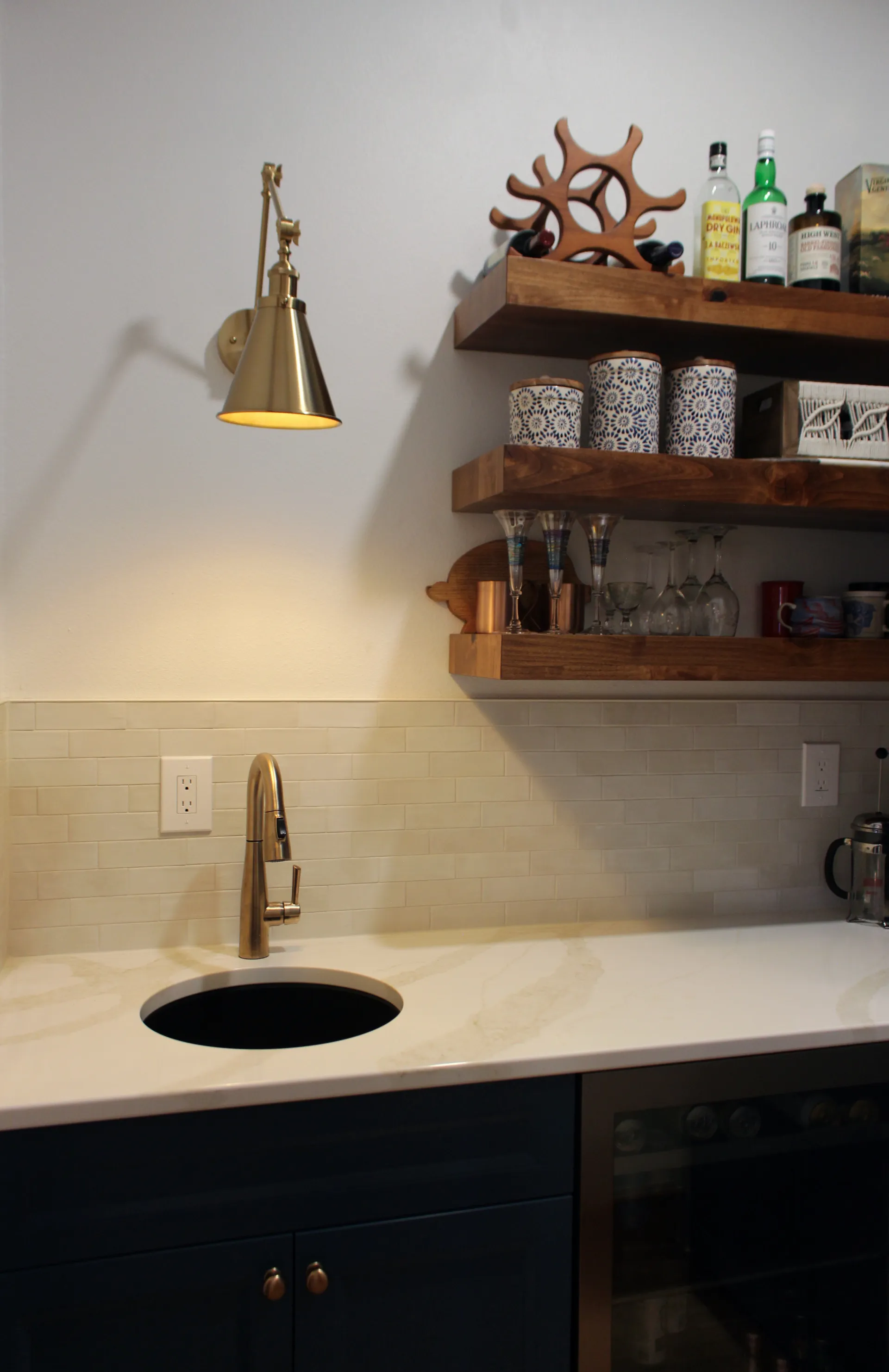 Modern kitchen with gold wall sconce, wooden shelves holding bottles and mugs, marble countertop, undermount sink, and tile backsplash. Warm ambiance.