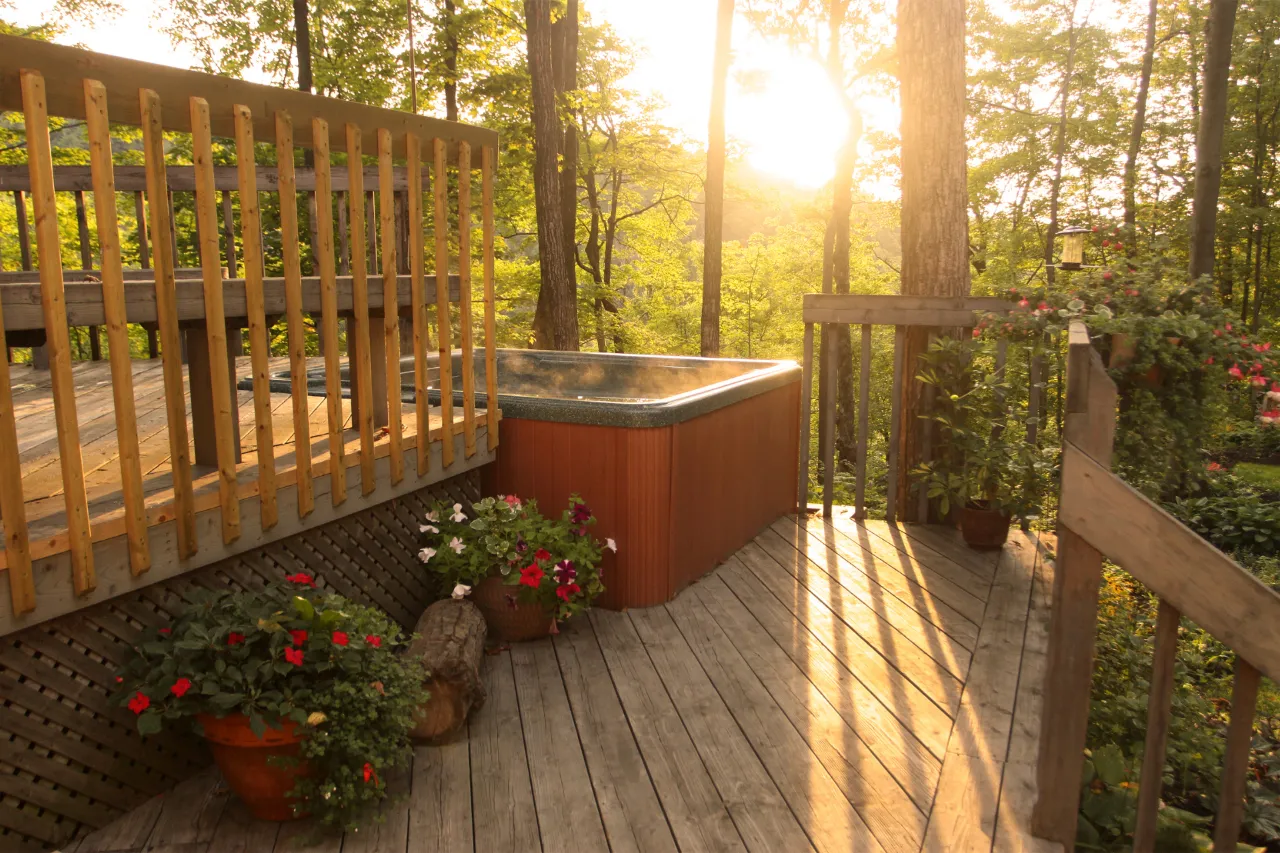 Sunlit wooden deck with a hot tub, surrounded by lush green trees. Potted flowers add color, creating a tranquil and inviting atmosphere.