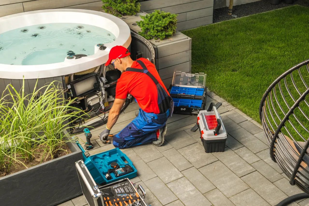 A technician in a red and blue uniform works on a spa pump outdoors. Toolboxes surround him on a patio. There’s greenery and a neatly manicured lawn.