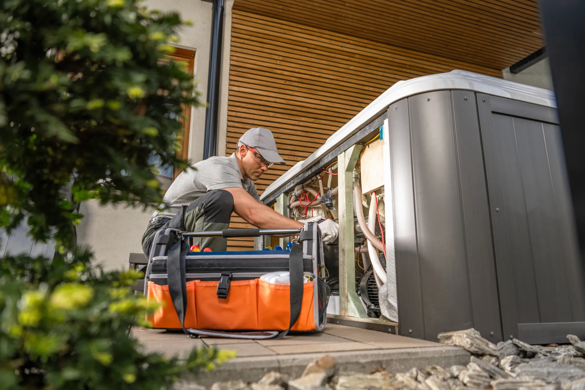 A technician in a cap and gray uniform kneels beside a hot tub, working on its exposed mechanical parts. An orange tool bag is in the foreground.