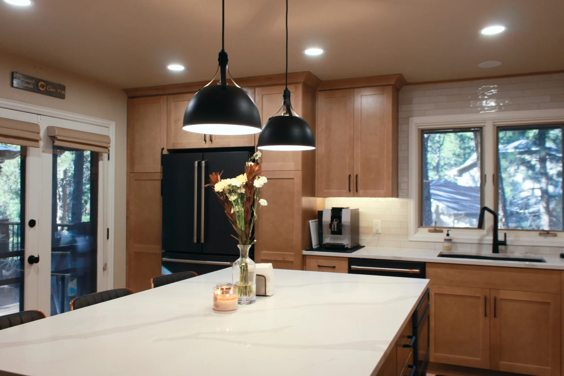Modern kitchen with wooden cabinetry, black fridge, and two pendant lights over a white island. A vase with flowers and candle add a cozy touch.