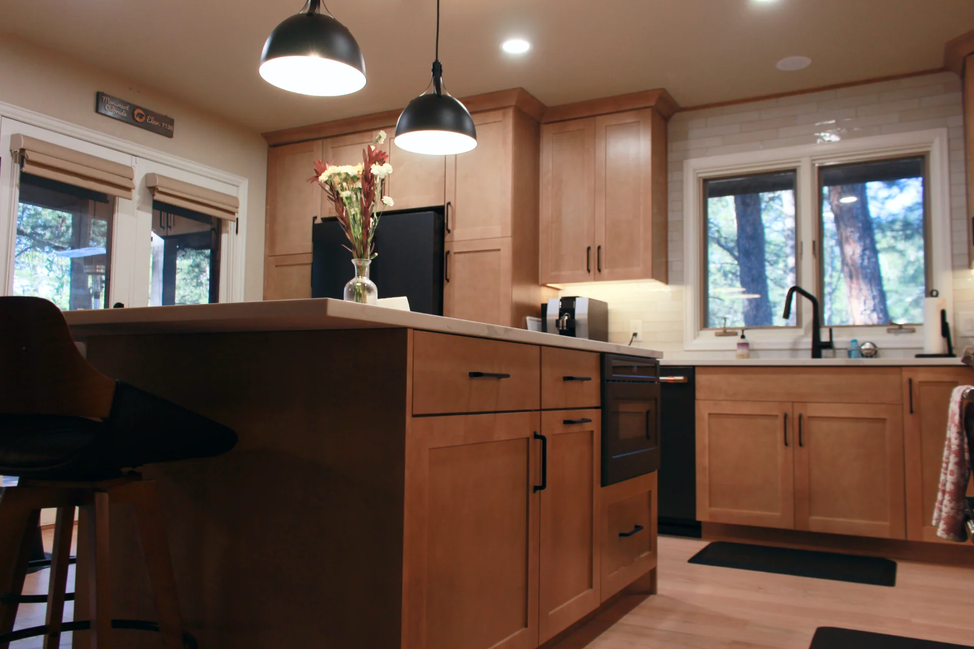 A cozy kitchen with warm wooden cabinets, black appliances, and soft lighting. A vase with flowers sits on the island beneath hanging lamps.