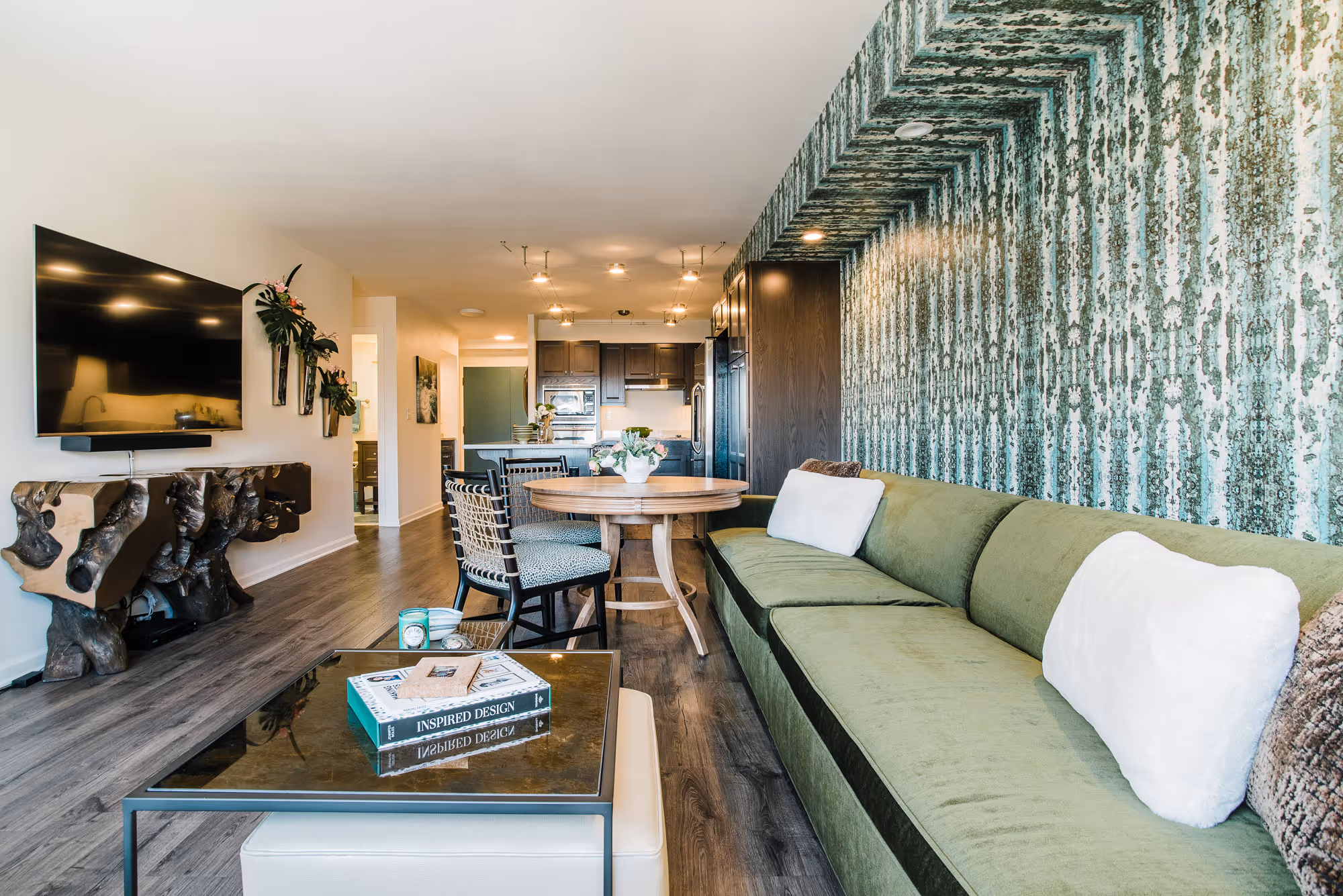 Modern living room interior design with a green couch, patterned accent wall, round wooden dining table, and mounted flat-screen TV.