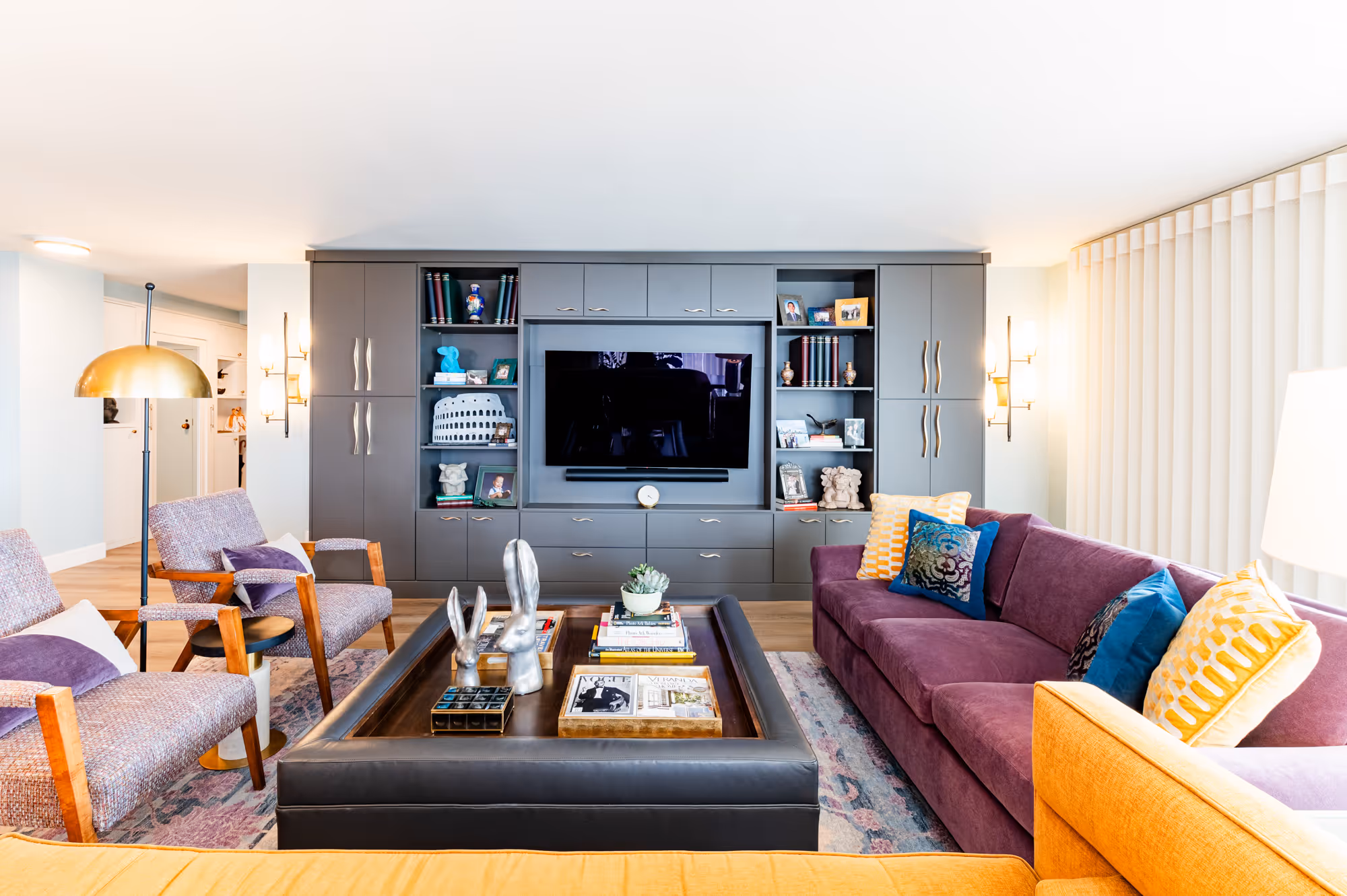 Modern living room with a large purple sofa, two patterned armchairs, a black ottoman coffee table with decorative items, and a gray entertainment unit with a flat-screen TV.