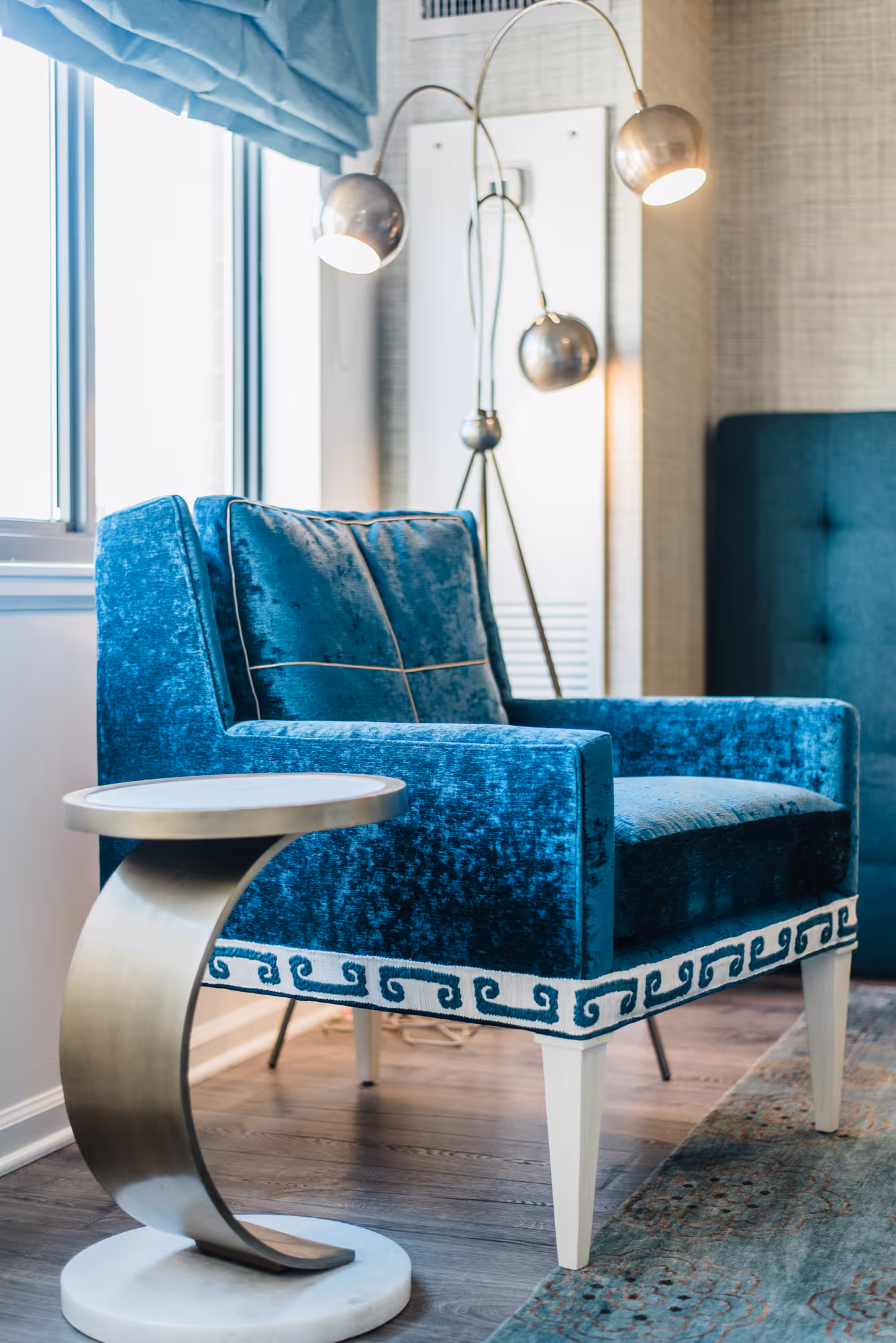 Blue velvet armchair with patterned trim next to a curved metallic side table by a window.