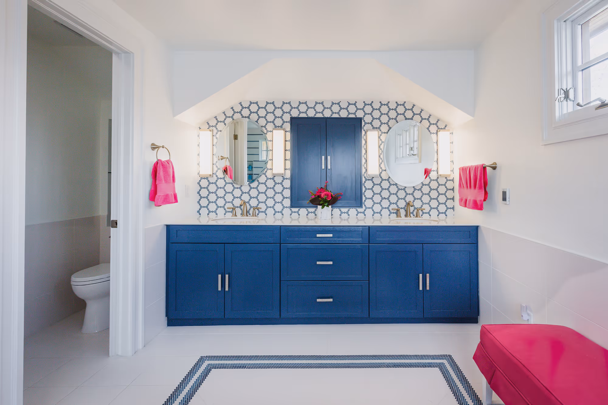 Bright bathroom remodel with blue double vanity, round mirrors, geometric blue and white backsplash, and pink accents including towels and bench.