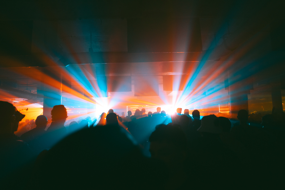 Silhouetted crowd at an indoor event with vibrant blue and orange stage lights radiating through haze.