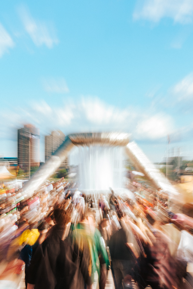 Crowd of people walking toward a large fountain in an outdoor urban area under a blue sky.