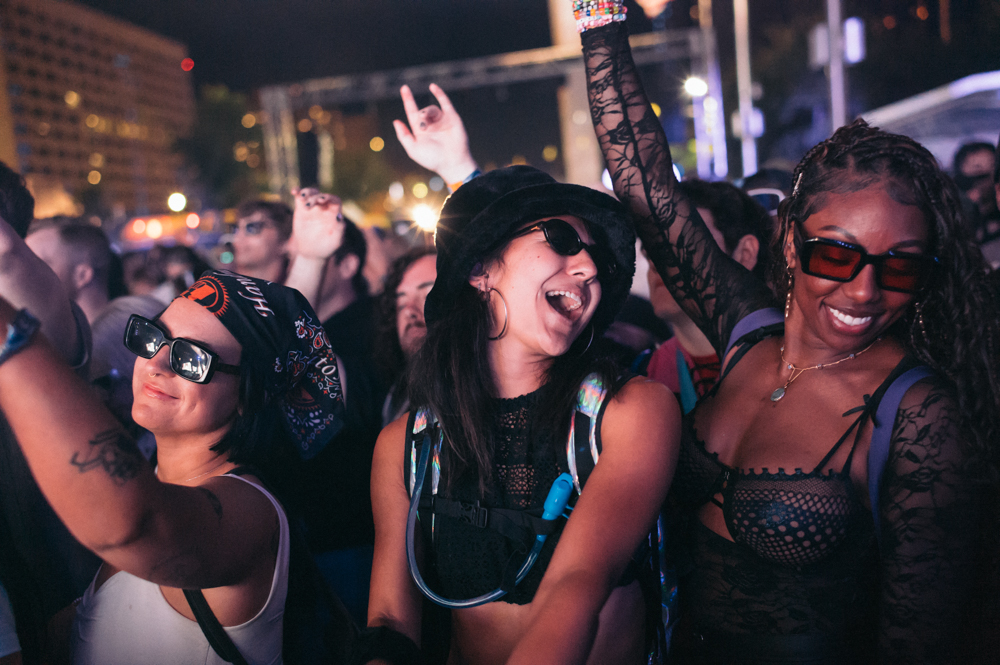 Three women wearing sunglasses happily dancing in a crowded outdoor nighttime event.