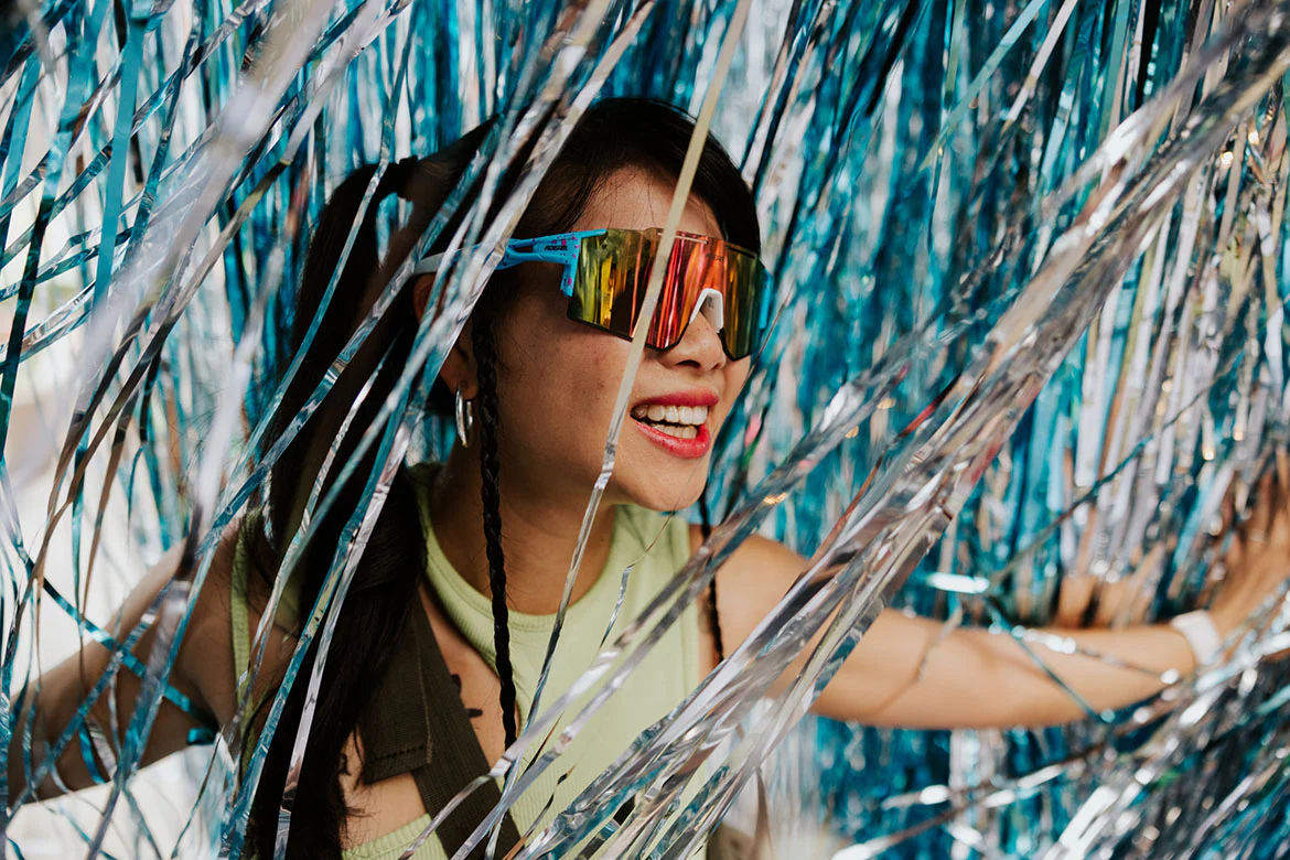 Smiling woman with braided hair wearing reflective sunglasses among blue and silver tinsel streamers.