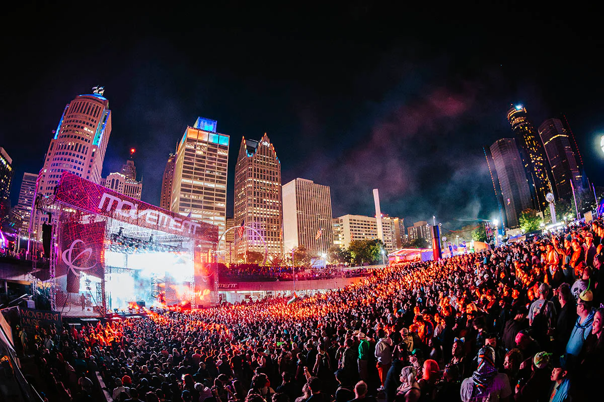 Large crowd attending an outdoor night concert with bright stage lights and city skyscrapers in the background.