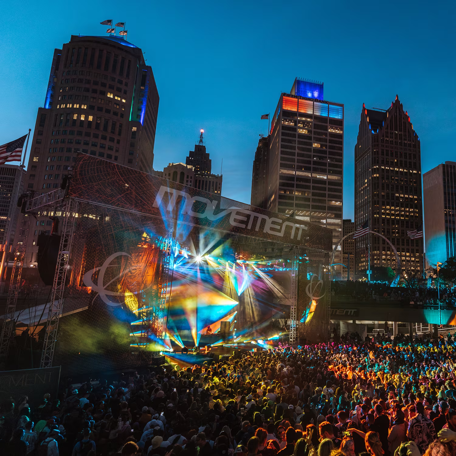 Large outdoor concert stage with colorful lights and a crowd in front, set against a city skyline at dusk.
