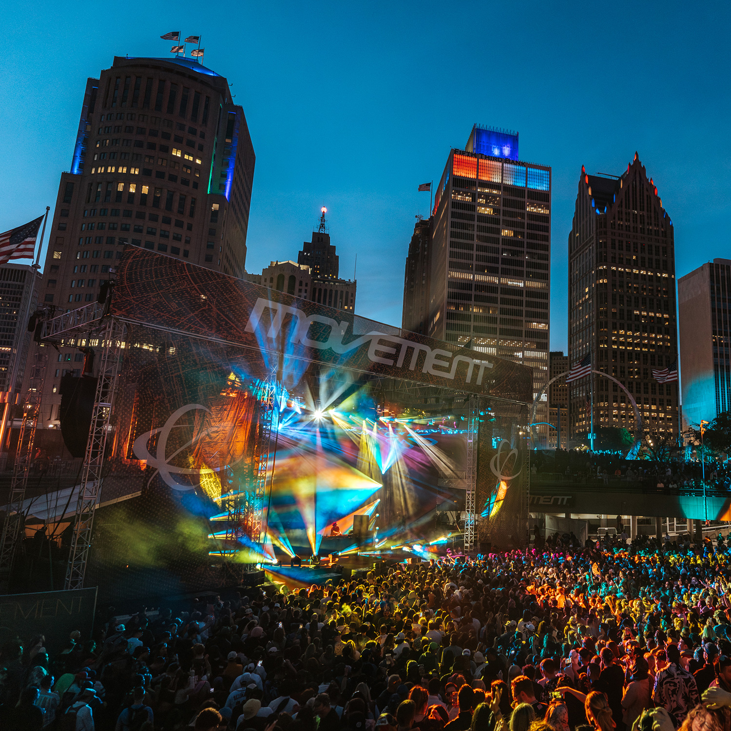 Large outdoor concert stage with colorful lights and a crowd in front, set against a city skyline at dusk.