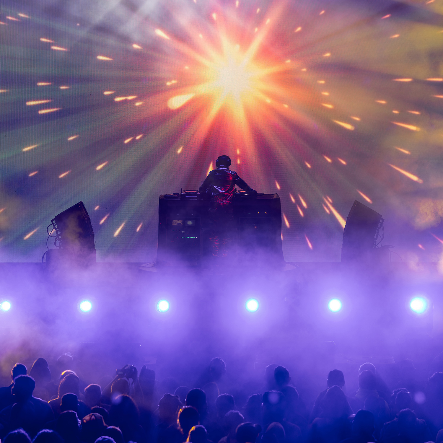 DJ performing on stage with bright colorful light effects and fog, facing a crowd at a live event.