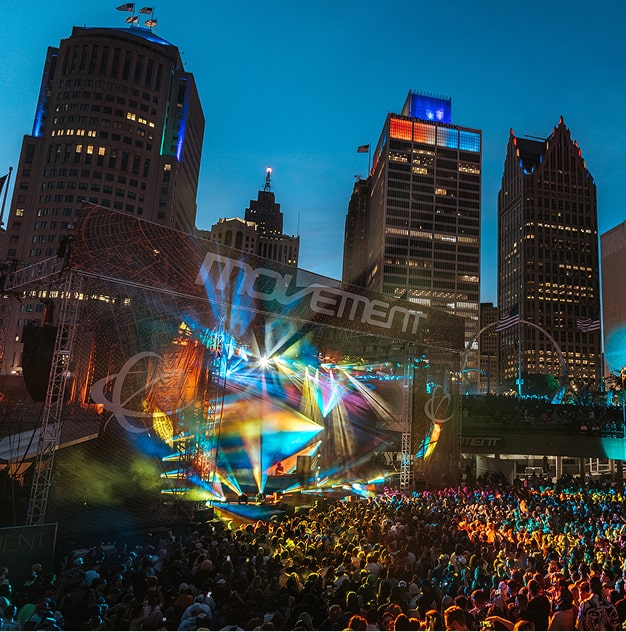 Large outdoor concert stage with colorful lights and a packed crowd in a cityscape at dusk.