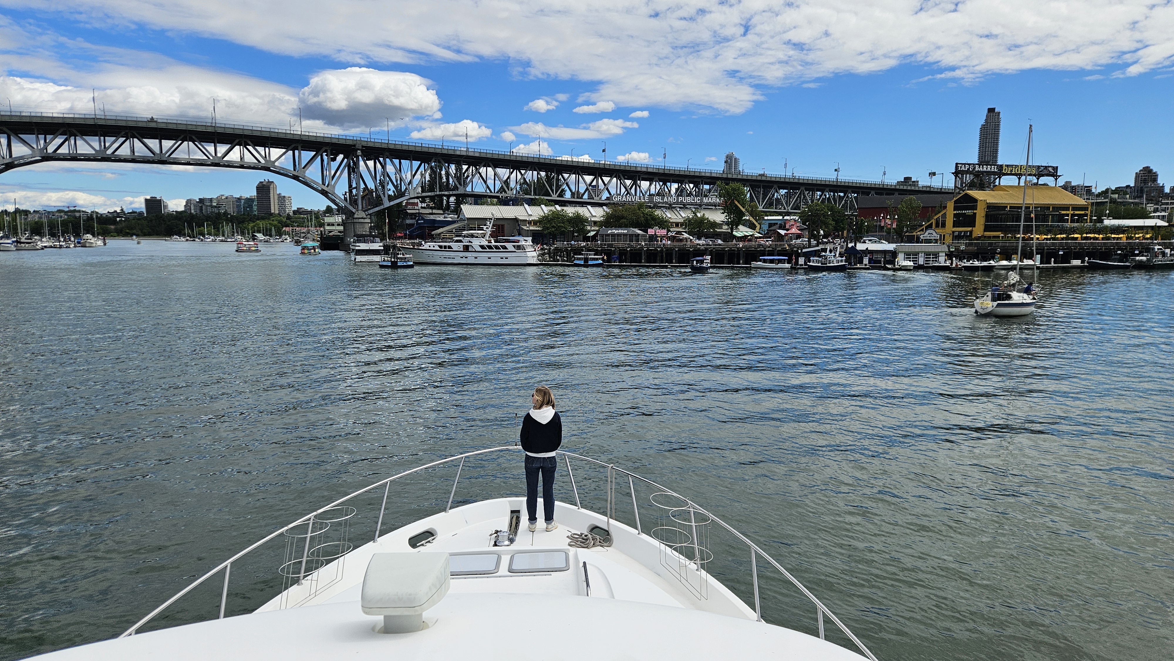 Person standing on a boat's bow looking toward a city waterfront with a bridge and boats.