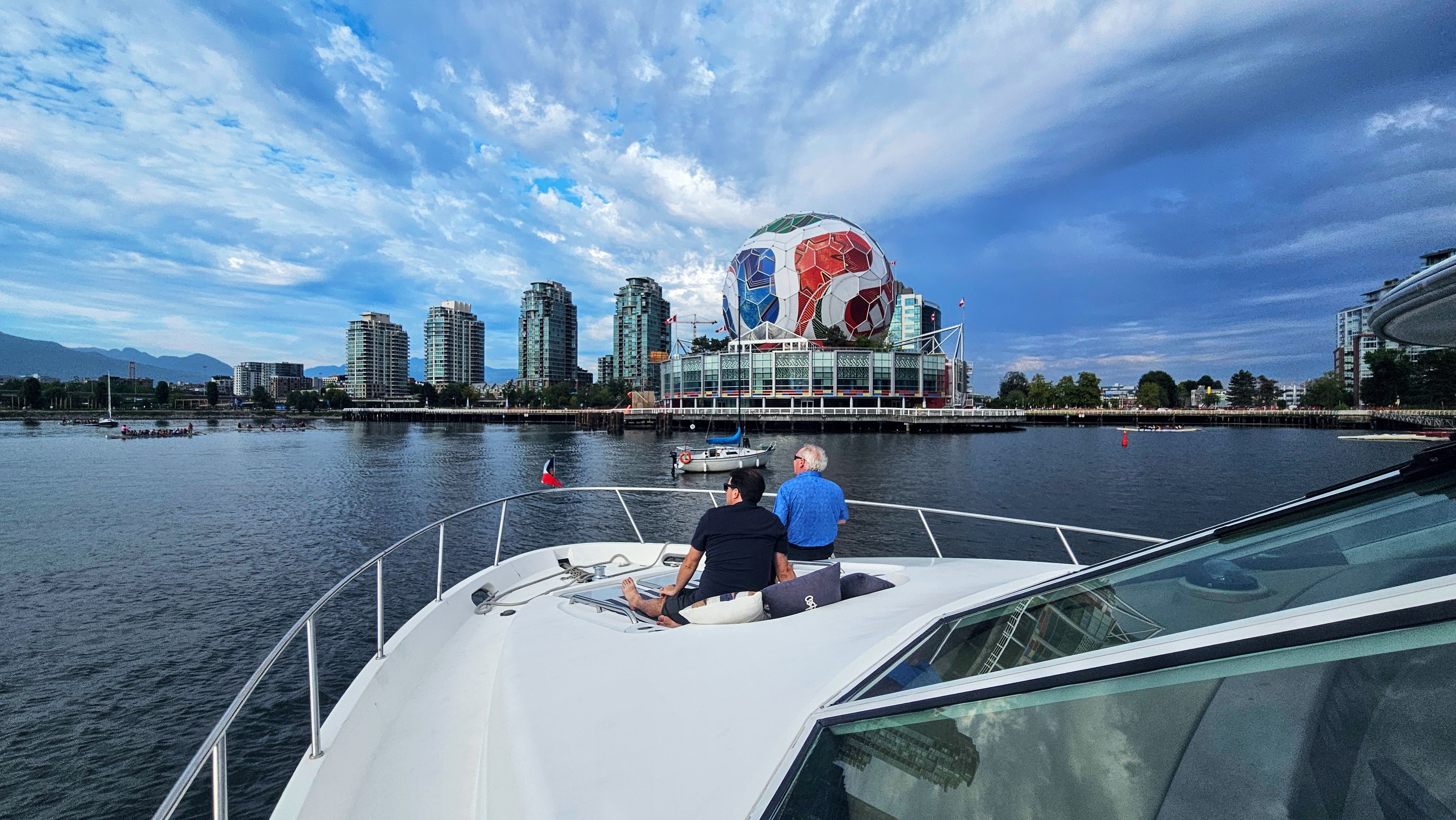 Person sitting on a yacht's bow looking toward city skyline and mountains across the water at sunset.
