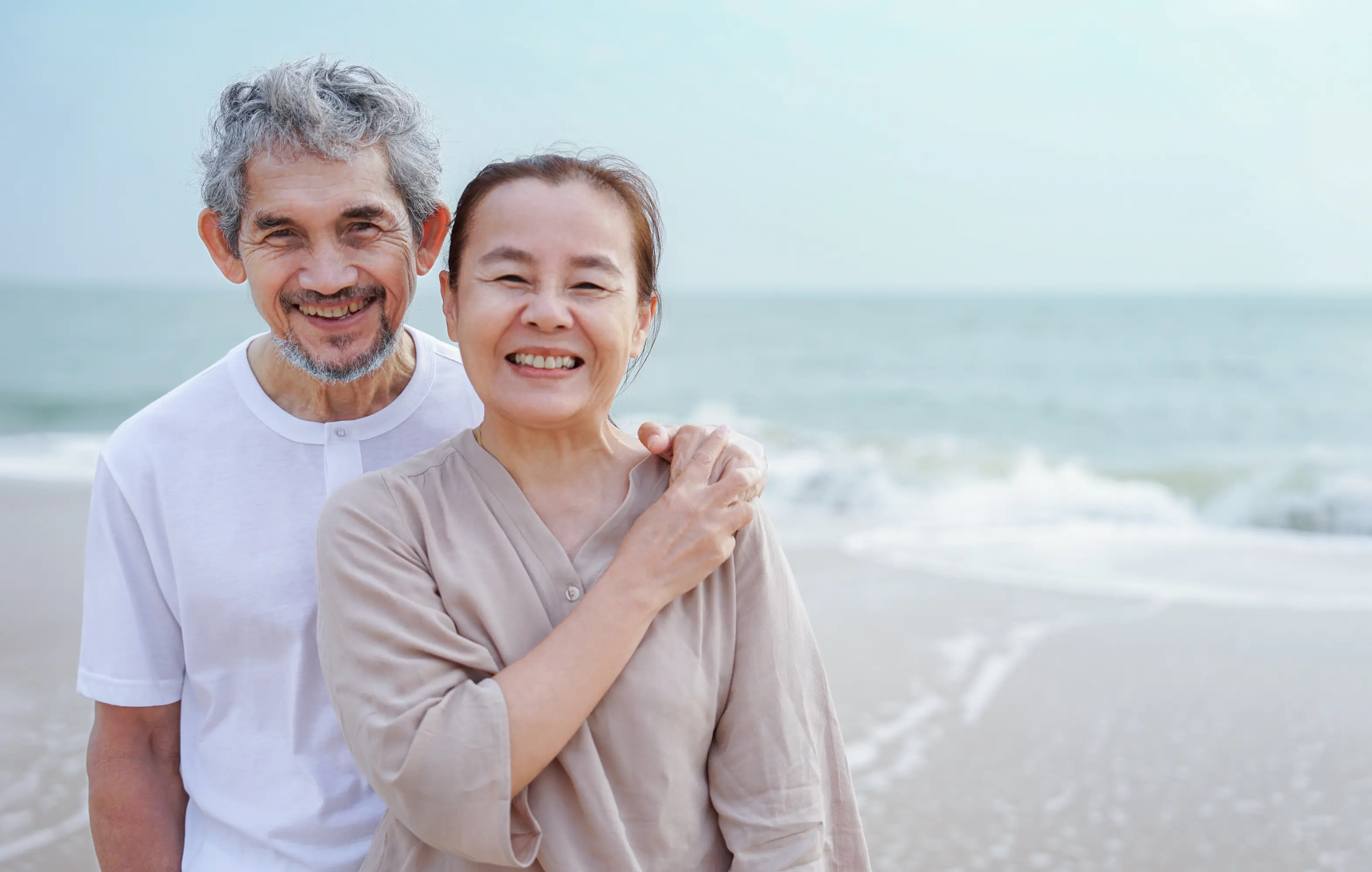 Smiling elderly couple standing on a beach with ocean waves in the background.
