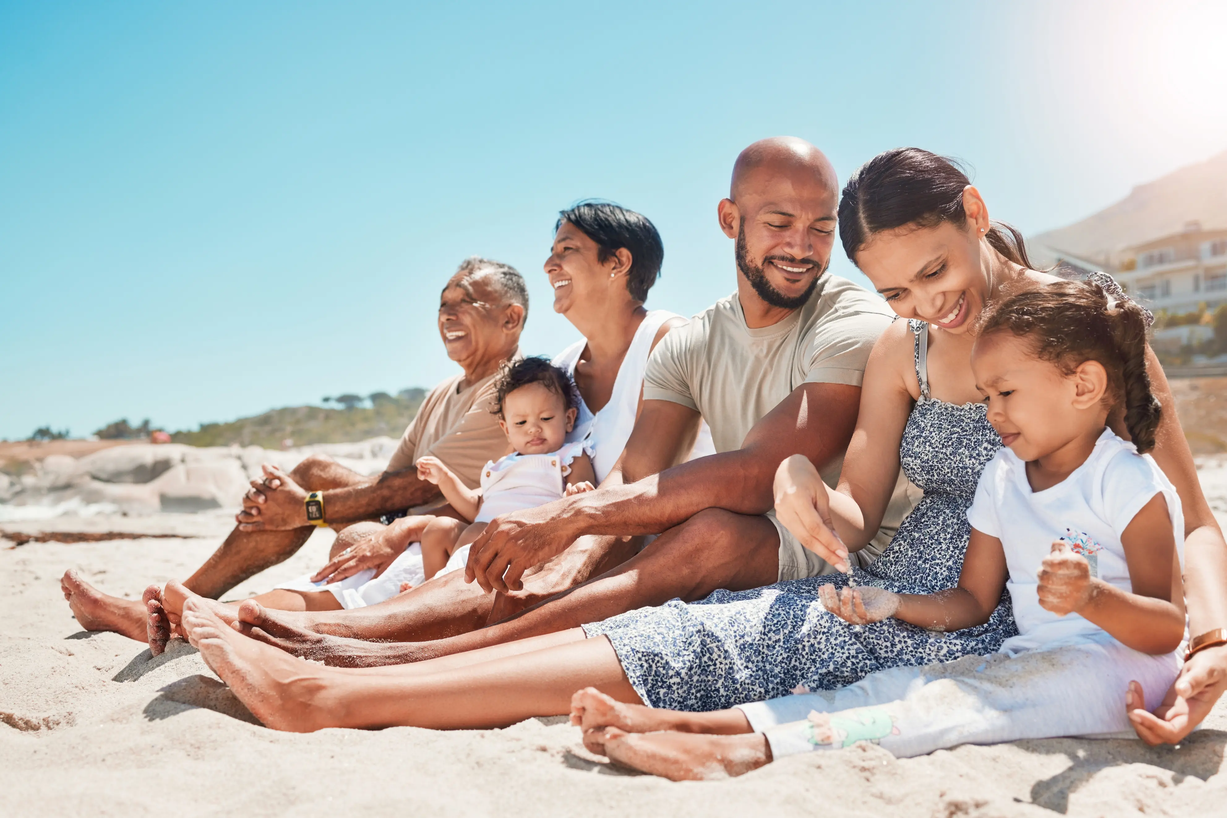 Multi-generational family sitting on the sand at the beach on a sunny day, smiling and enjoying each other's company.