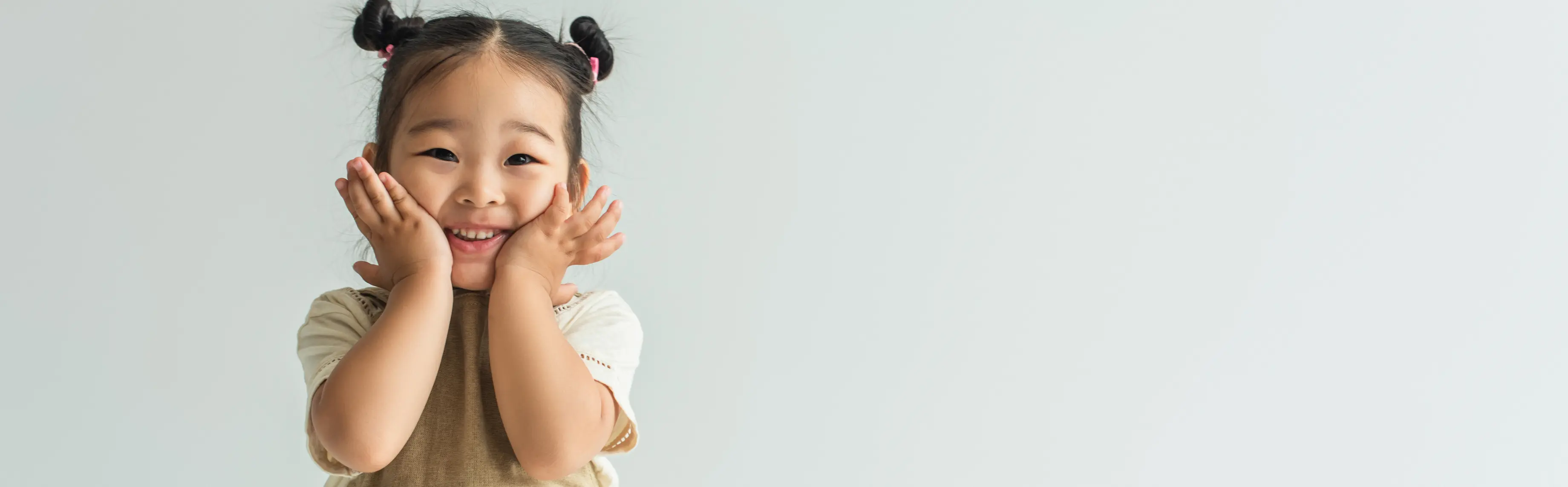 Smiling young girl with two buns holding her face with both hands against a plain light background.