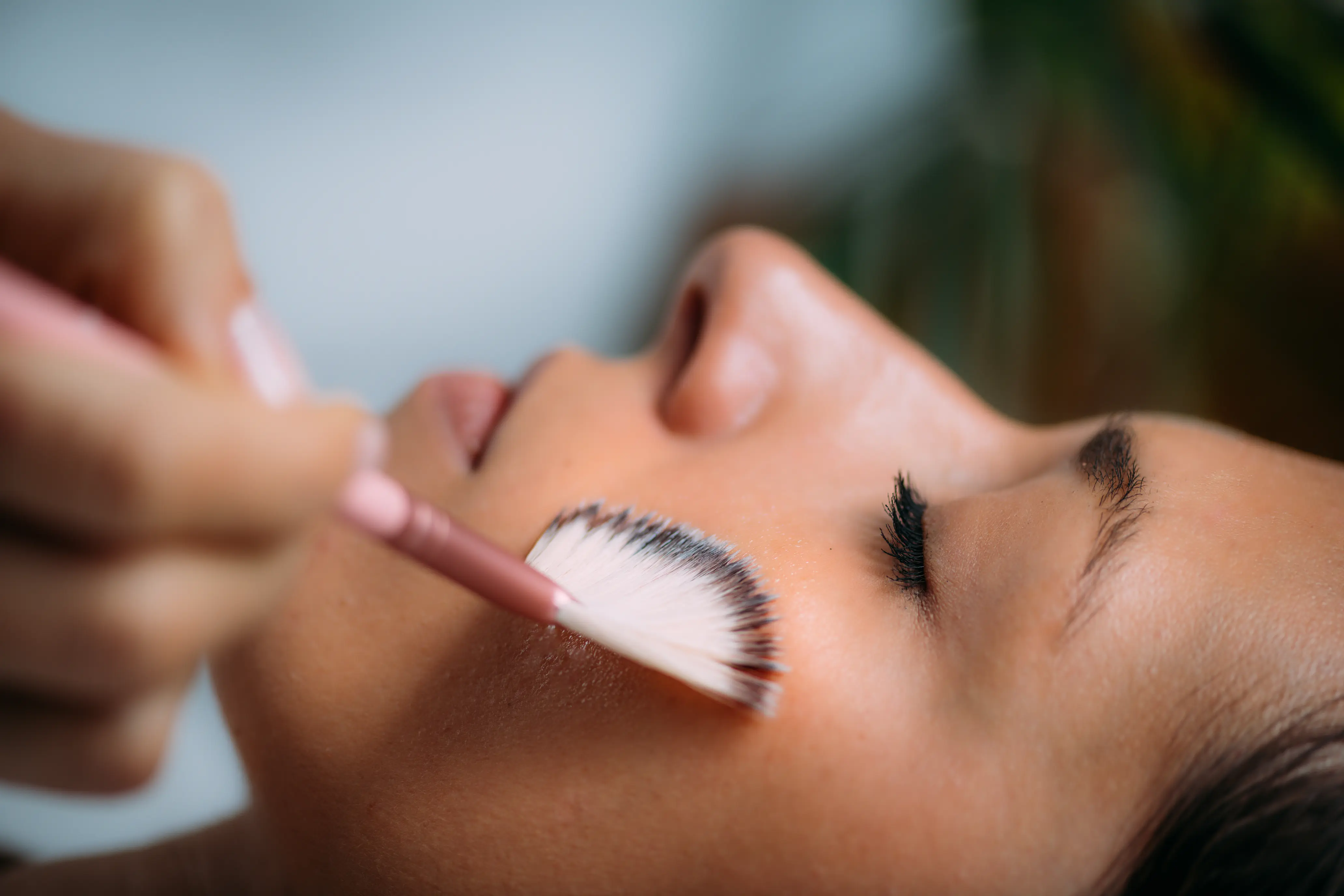 Close-up of a woman with eyes closed applying makeup to her cheek with a fan brush.