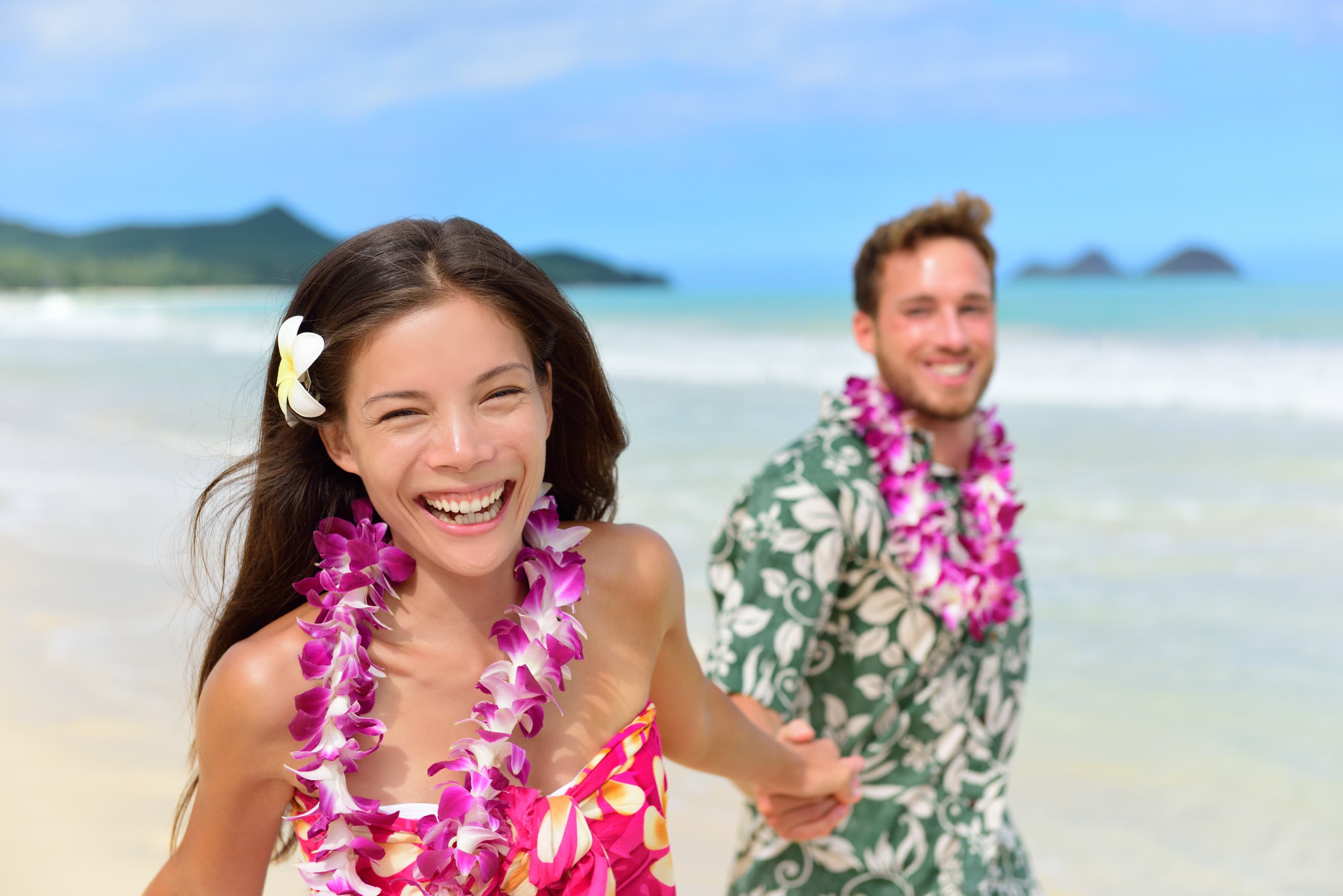 Smiling couple wearing floral leis holding hands on a sunny beach with ocean and islands in the background.