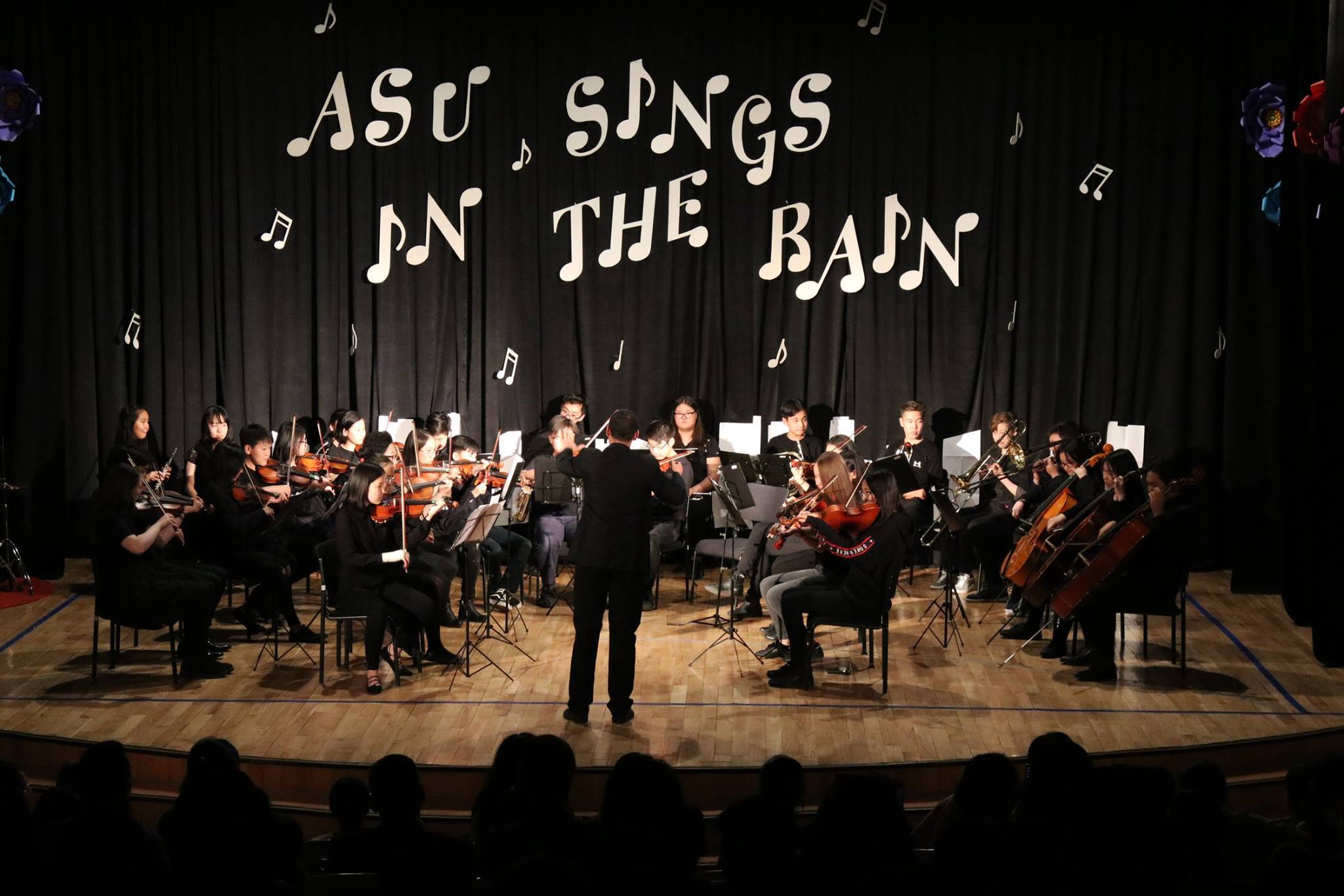 Orchestra performing on stage with conductor in front, backdrop reads 'ASU Sings in the Rain' with musical notes.