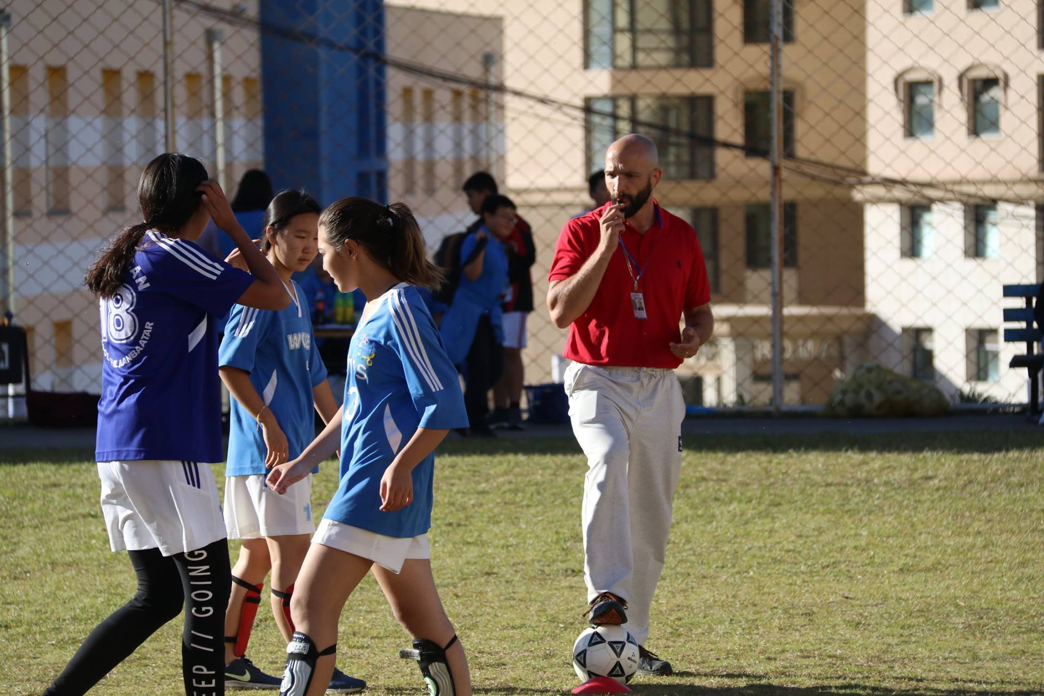 A coach in a red shirt and gray pants blowing a whistle while standing with a soccer ball on a grassy field near three female soccer players in blue jerseys.