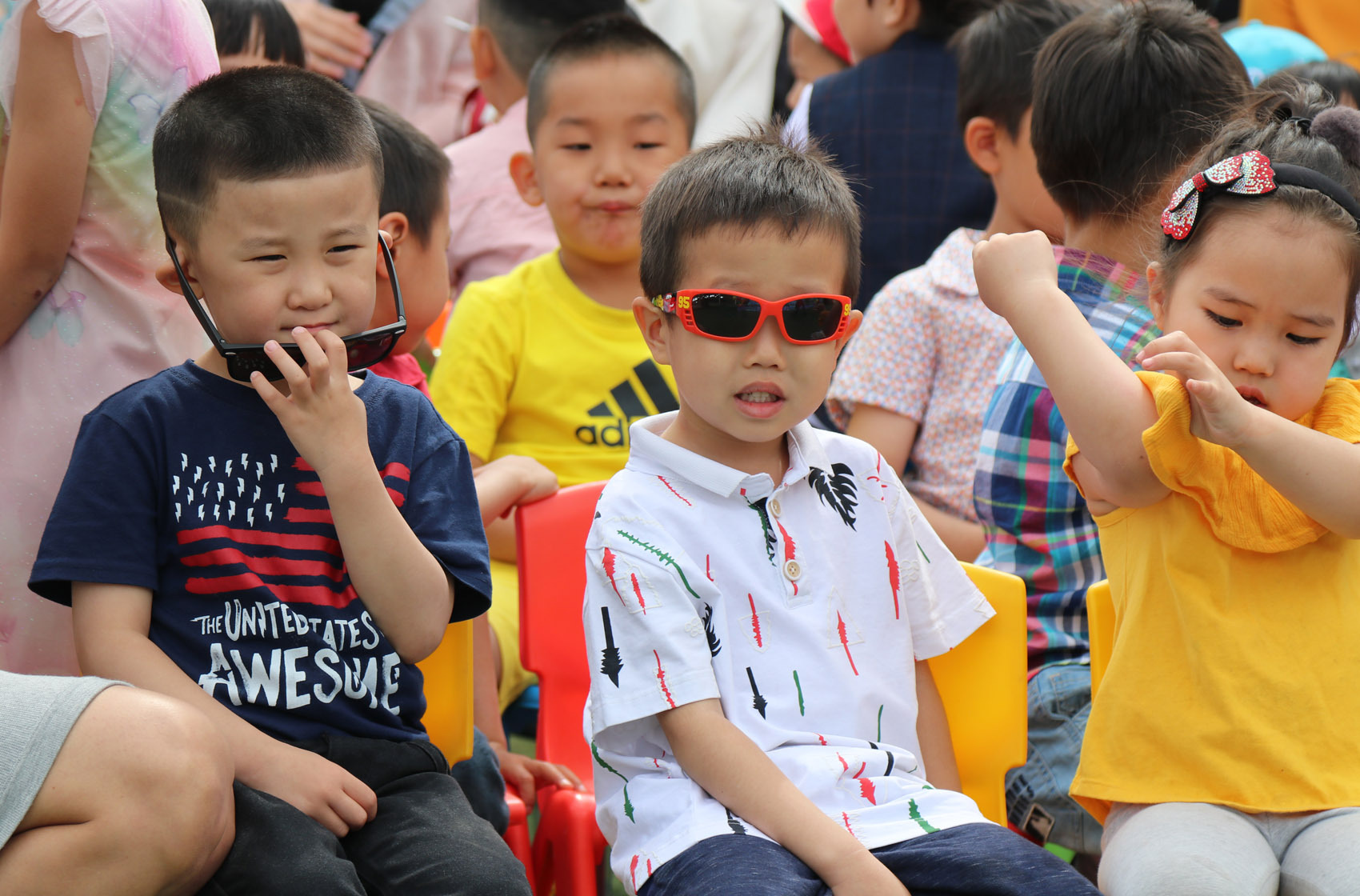 Group of young children sitting, one boy wearing red sunglasses and a white patterned shirt, another boy holding sunglasses near his face, and a girl in a yellow top looking at her arm.