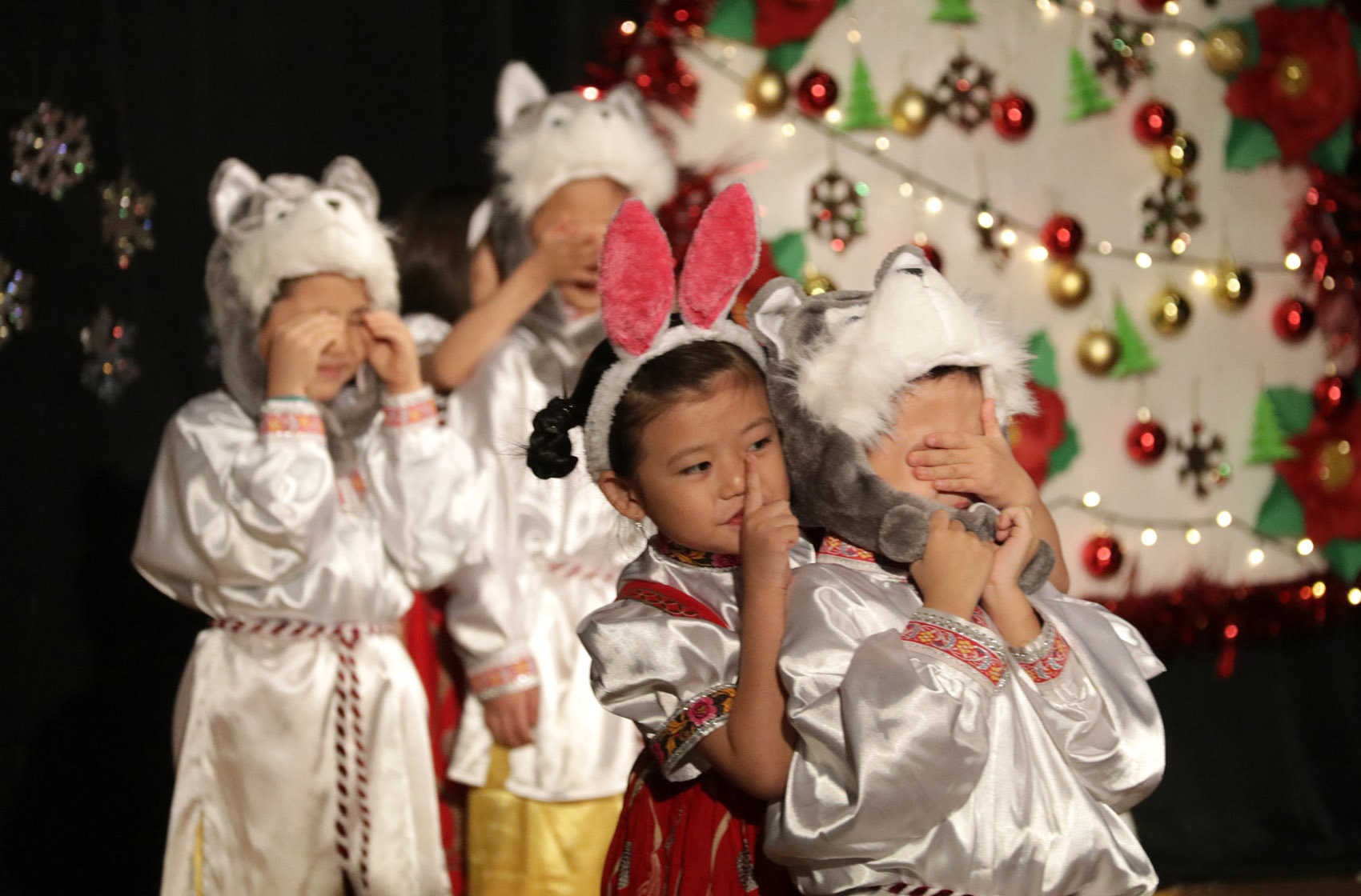 Children in animal costumes and festive dress performing on stage with holiday decorations in the background.
