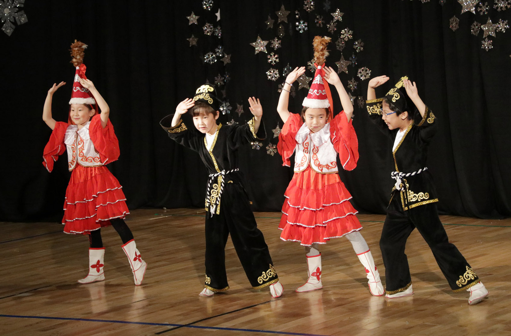 Four children performing a traditional dance in colorful costumes with red and black outfits and decorative hats on a wooden stage.