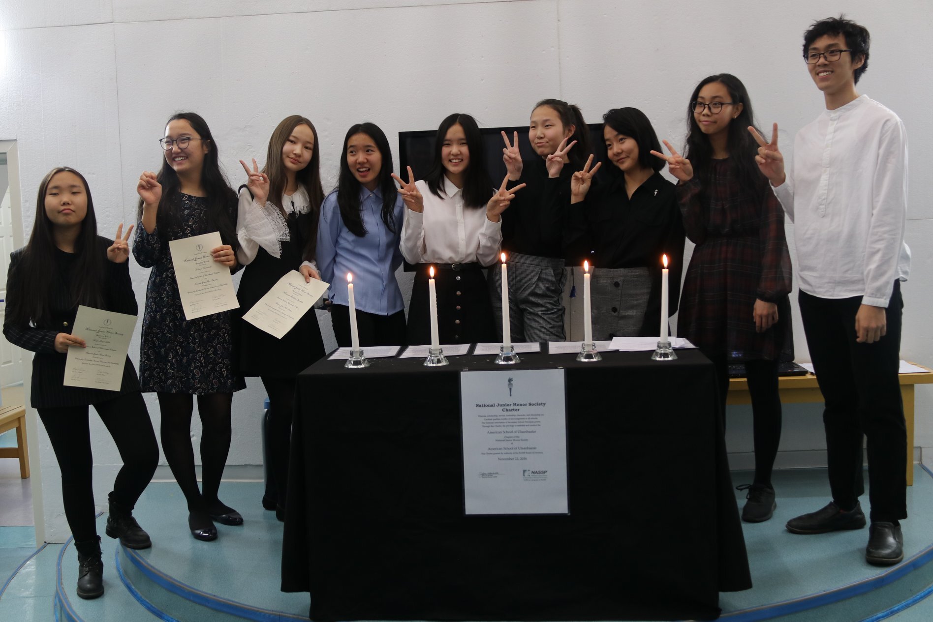Group of nine young people standing behind a table with five lit candles and certificates, all making peace signs.