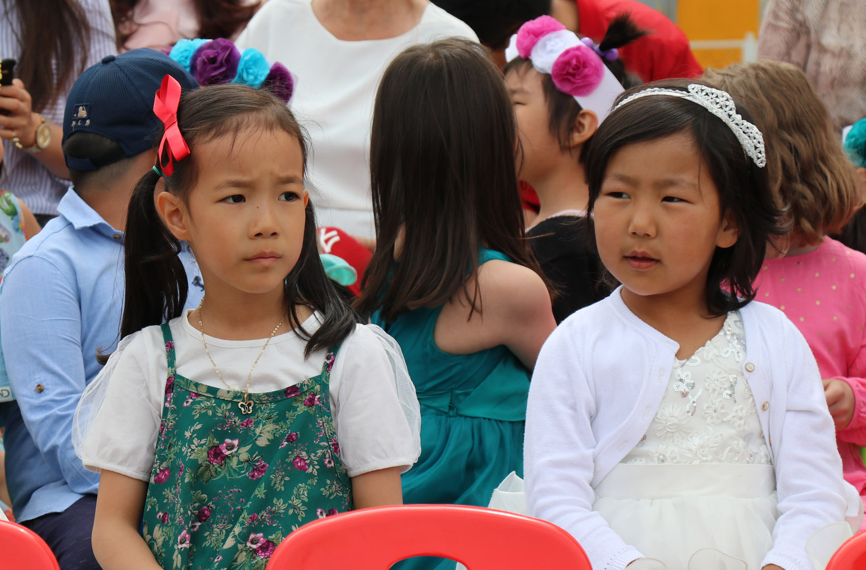 Two young girls sitting on red chairs, one with a green floral dress and red hair bow, the other in a white dress with a white headband.