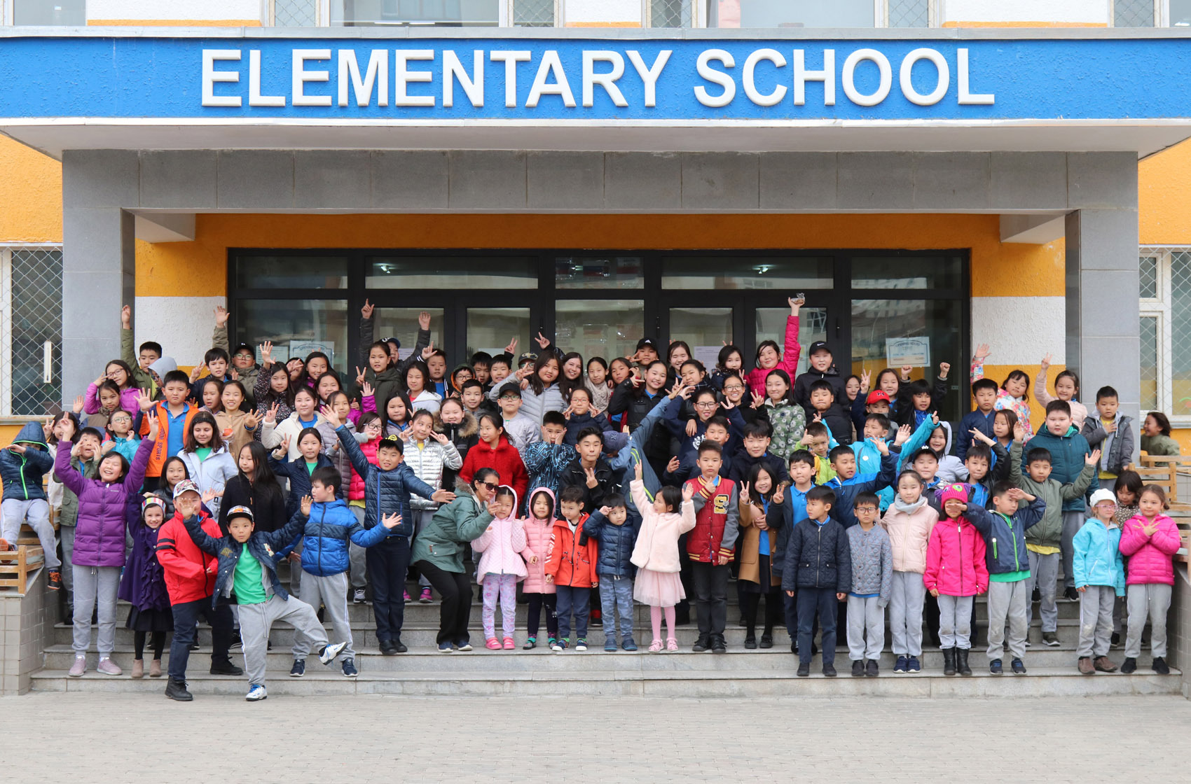 Large group of elementary school children and a teacher posing on the steps in front of a school building with a sign that reads 'ELEMENTARY SCHOOL'.
