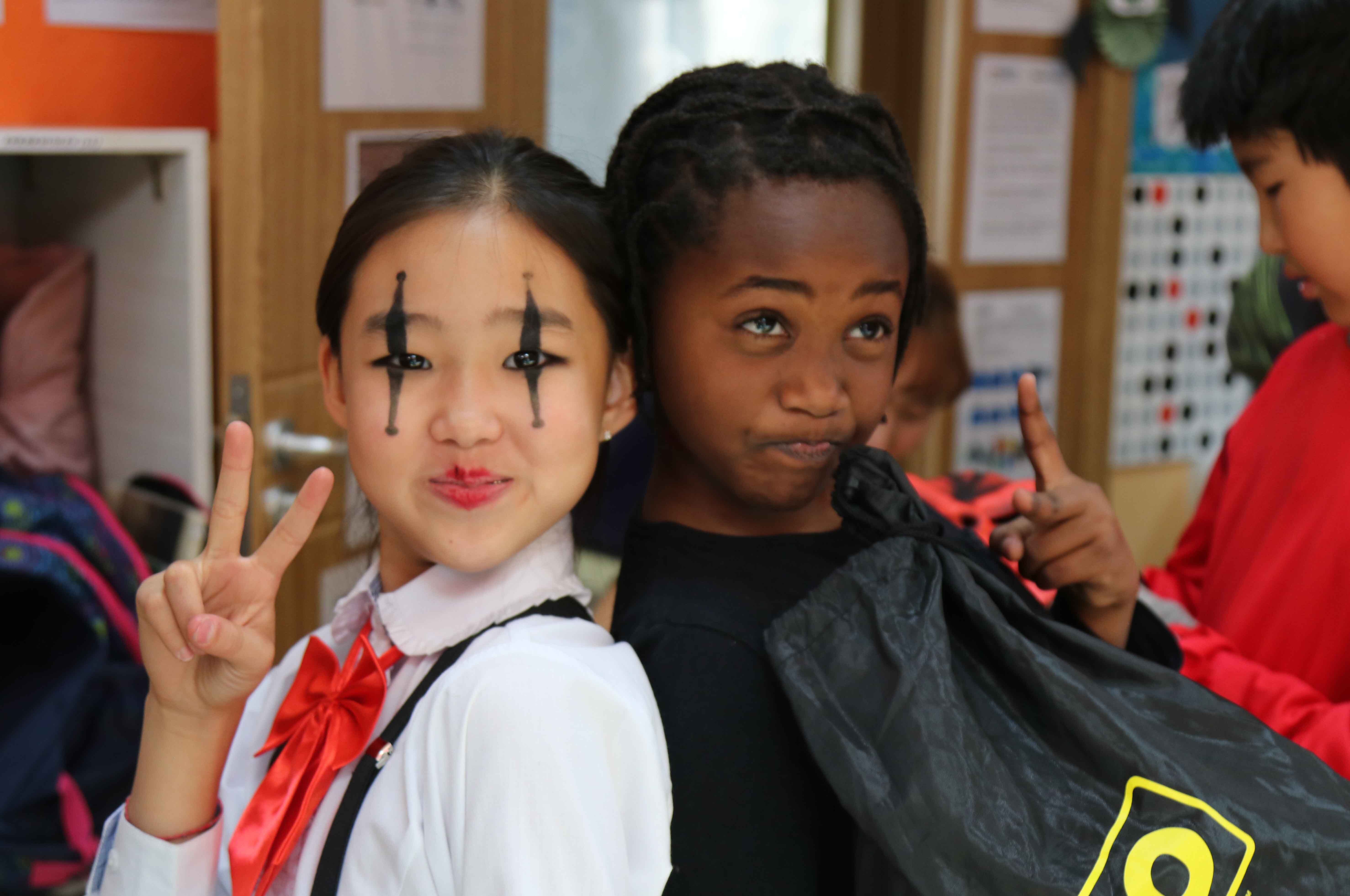 Two children posing with peace signs; one with black face paint resembling clown makeup and a red bow tie, the other with braids holding a black bag.