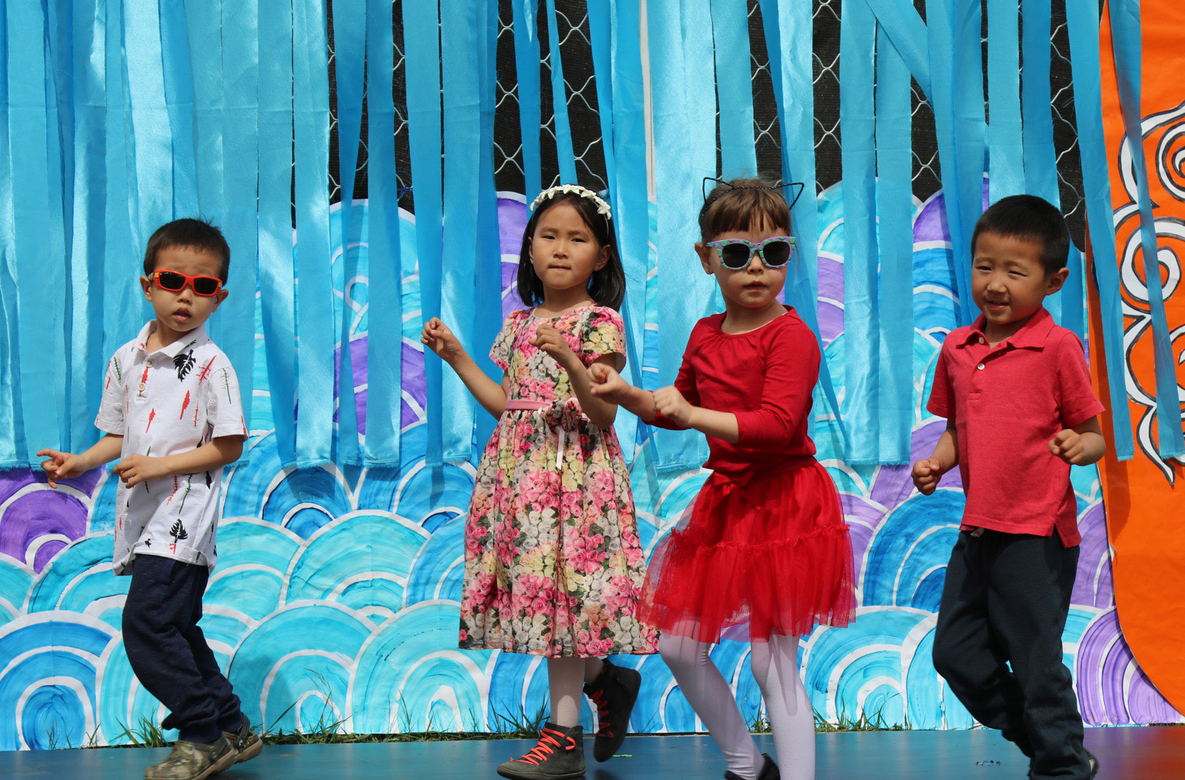 Four young children dancing on a stage with a colorful blue and purple backdrop, two girls in dresses and two boys in casual clothes.