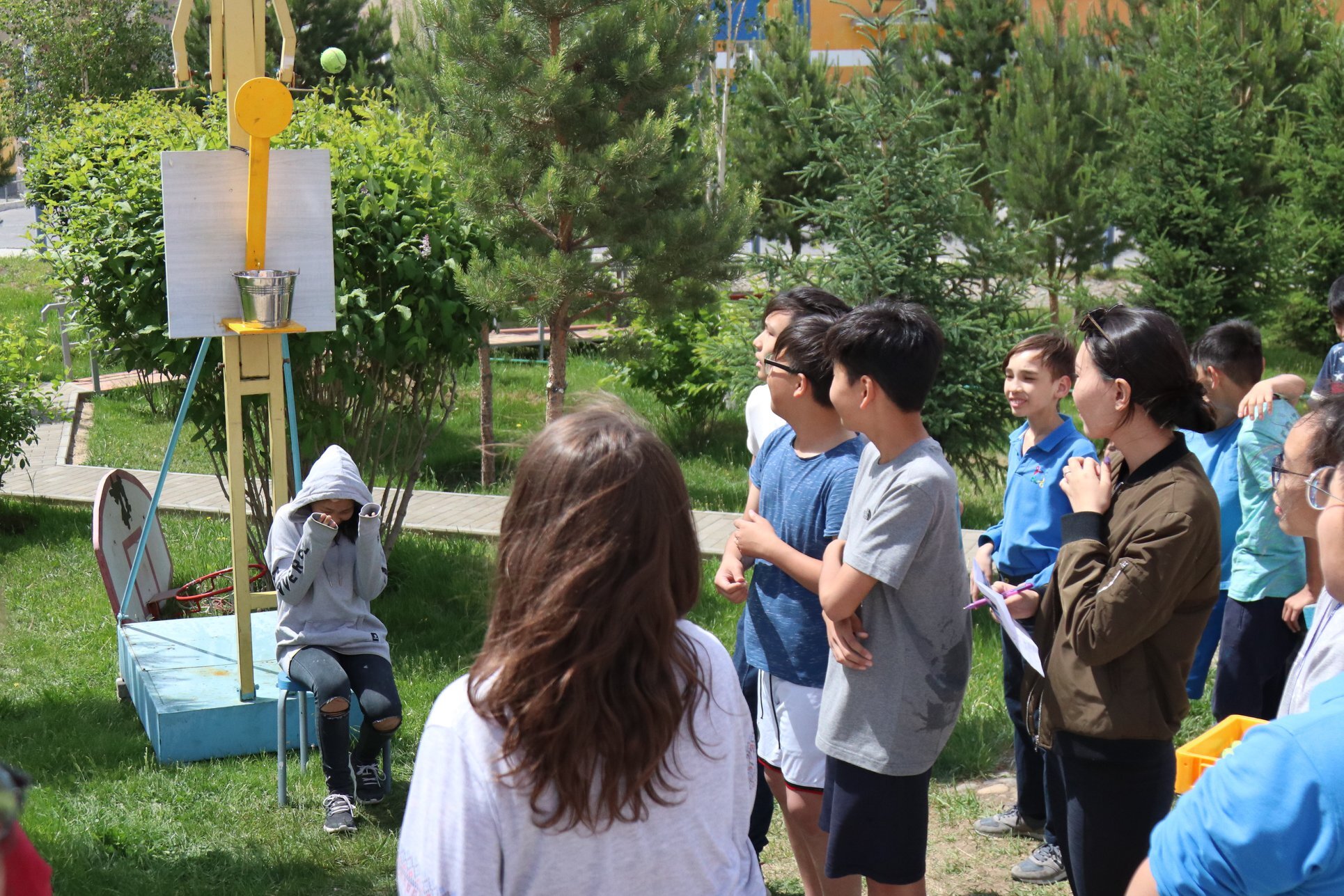 A group of children outdoors watching a girl in a gray hoodie sitting on a chair with her hands raised near her face, while a tennis ball flies toward a bucket mounted on a yellow contraption.
