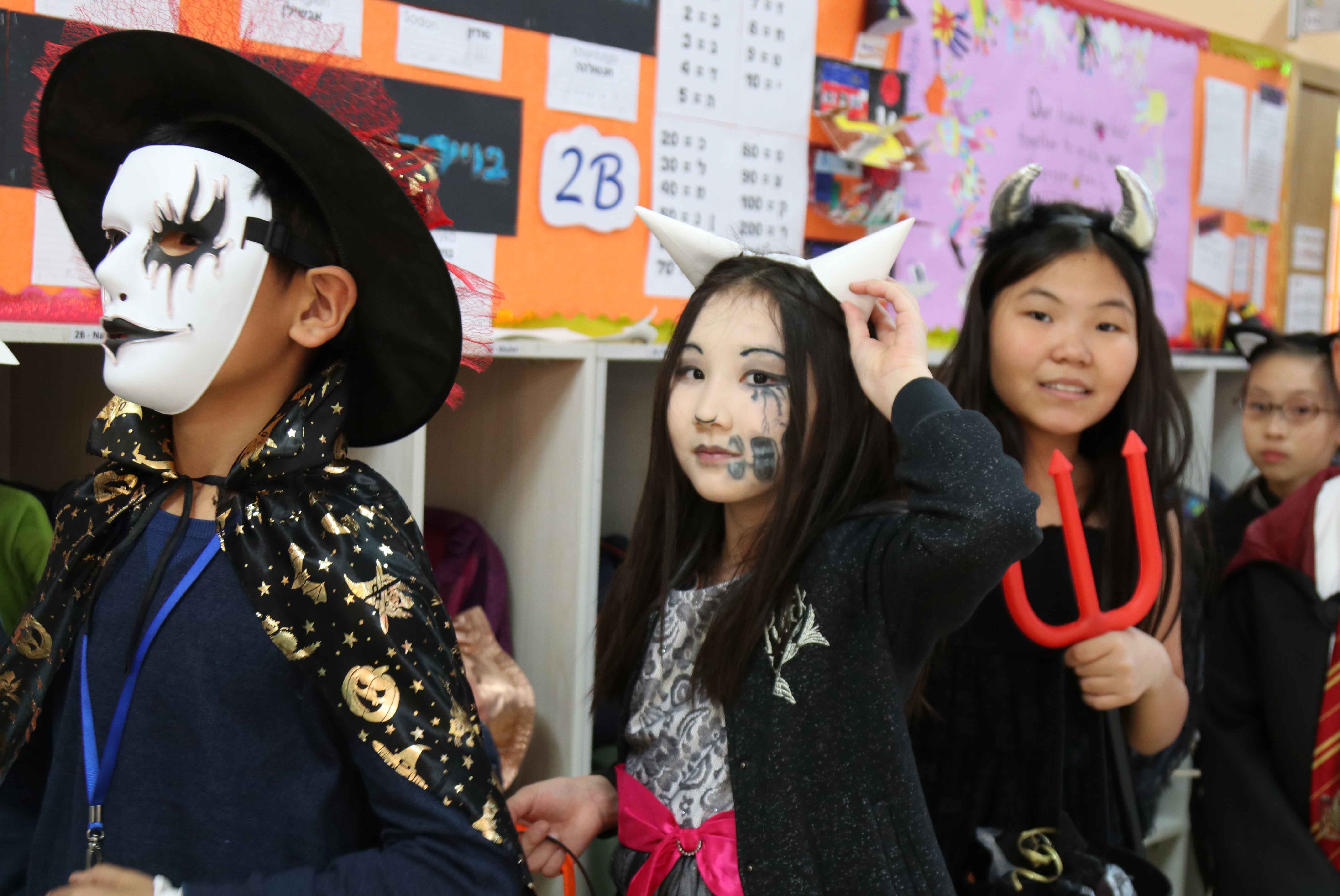 Children in Halloween costumes lined up indoors, including a child in a white mask and star-patterned cape, a girl with horns and face paint, and another girl with devil horns holding a red trident.