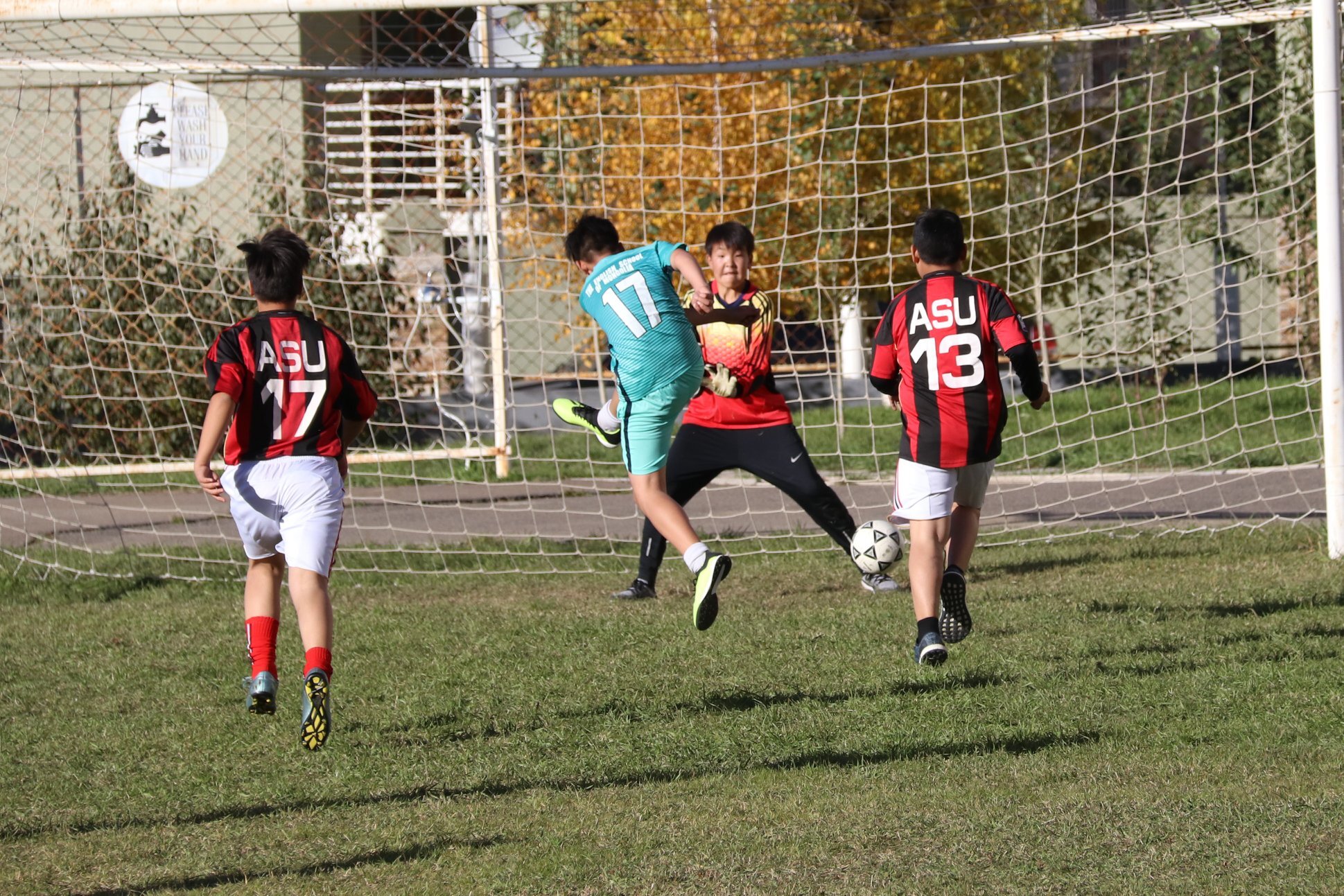 Youth soccer player in turquoise jersey kicks ball toward goal as goalkeeper and two defenders in red and black jerseys try to block.