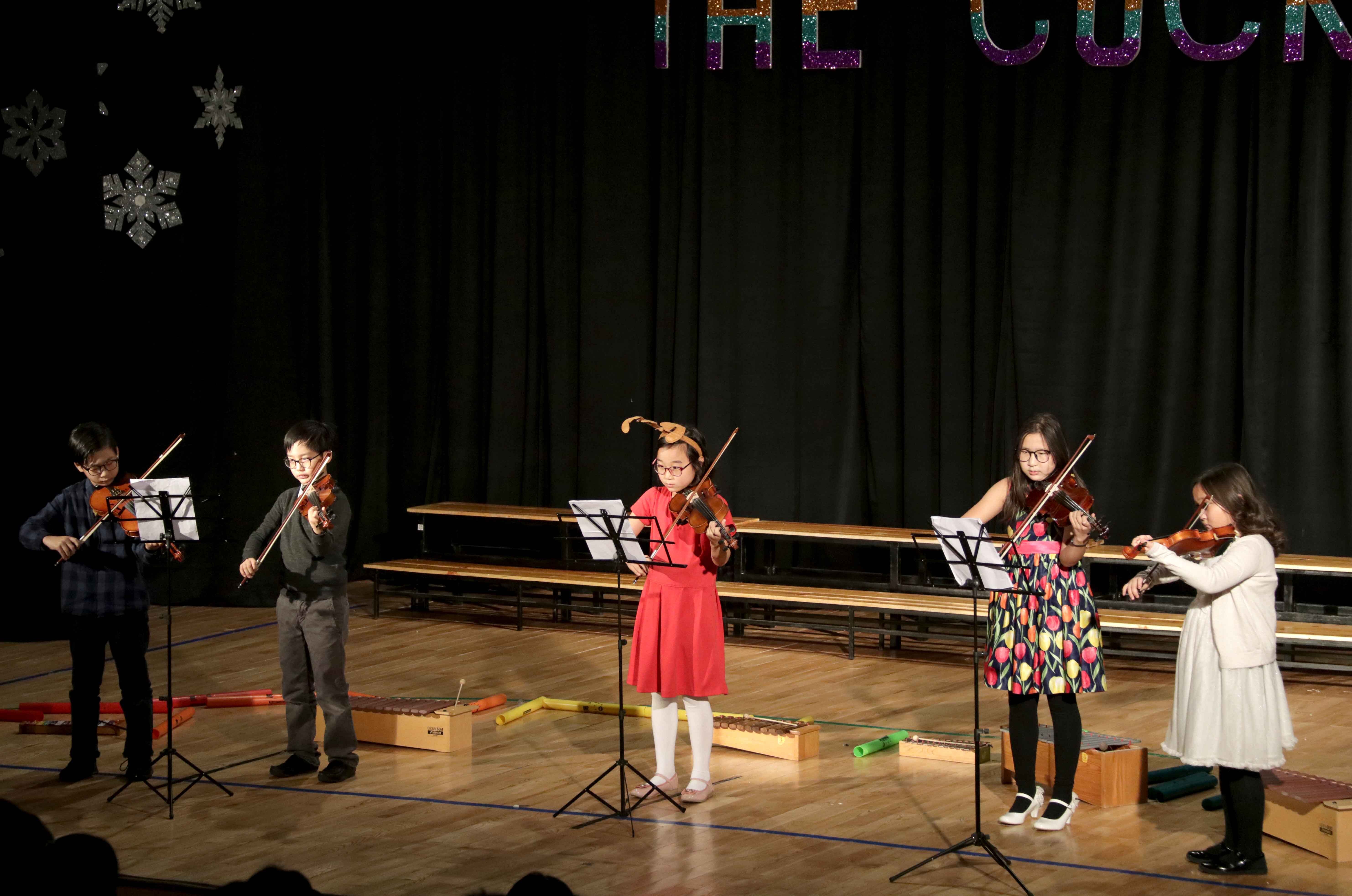 Five children playing violins on a stage with music stands and black curtains in the background.