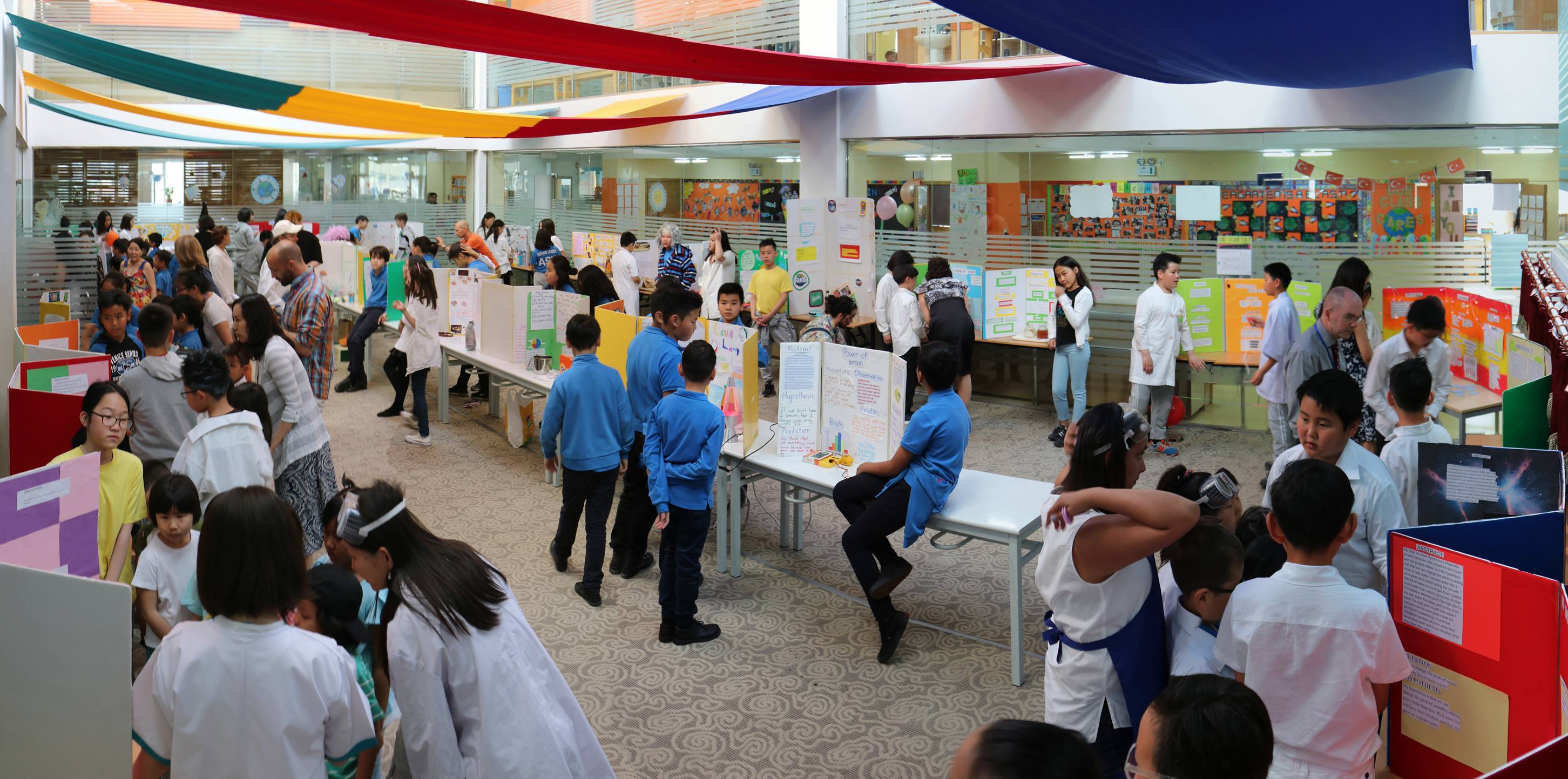 Children and adults gathered in a large indoor space for a science fair with colorful project boards displayed on tables.