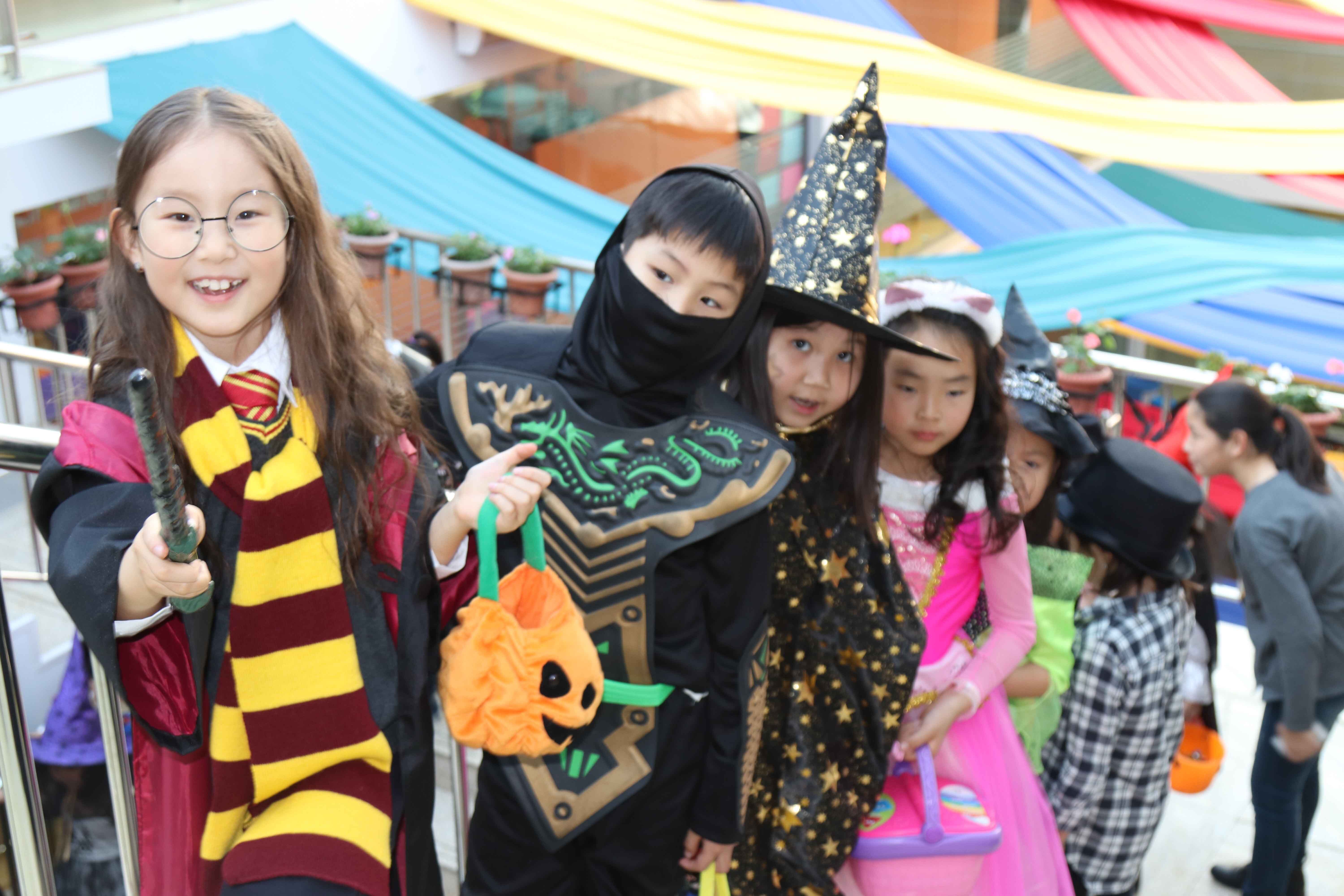 Children dressed in Halloween costumes including a wizard with glasses and wand, a ninja, a witch with a starry hat, and a princess carrying pumpkin bags.