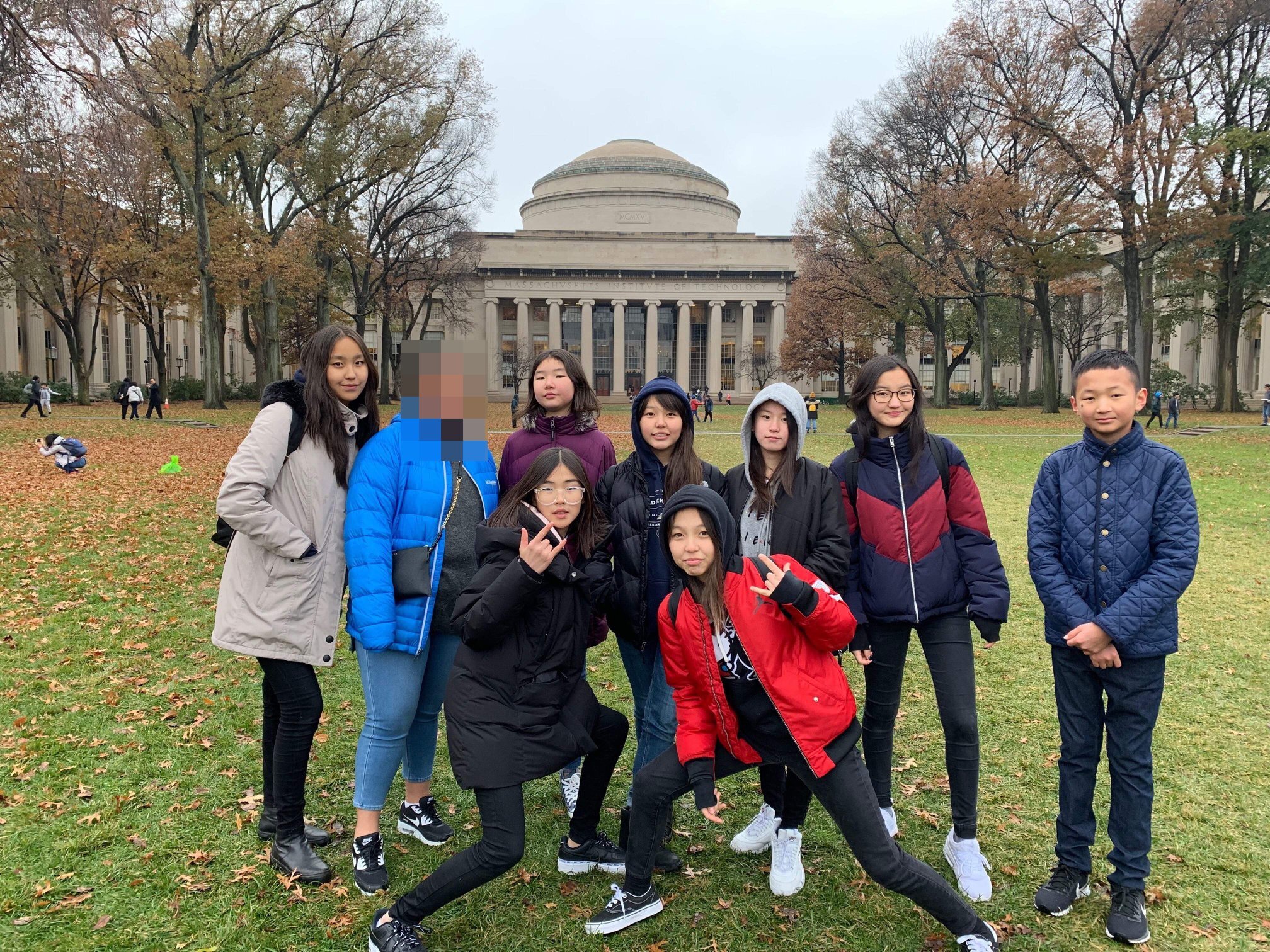 Group of eight young people posing outdoors on grass in front of a large building with columns and a dome, with fall trees around.