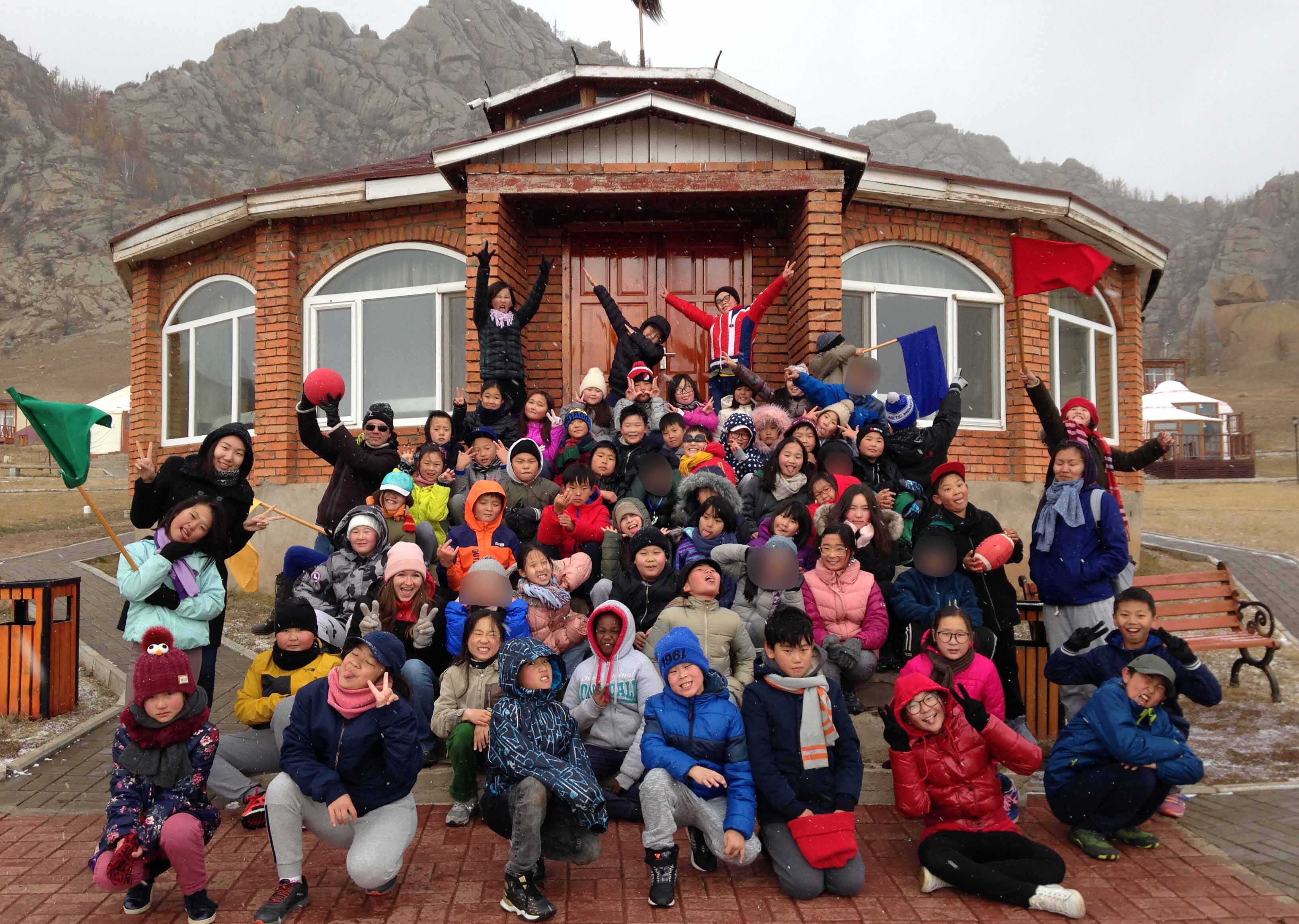 Large group of children and adults posing smiling in front of a brick building with mountains in the background, some holding colorful flags and a red ball.