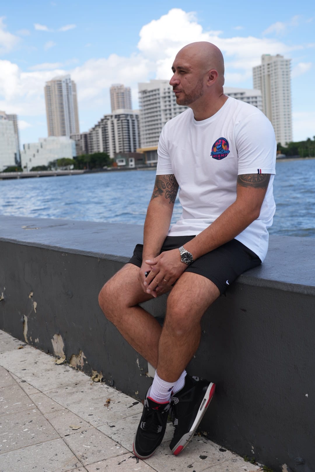 Man with tattoos wearing a white t-shirt and black shorts sitting on a concrete ledge by the water with city buildings in the background.