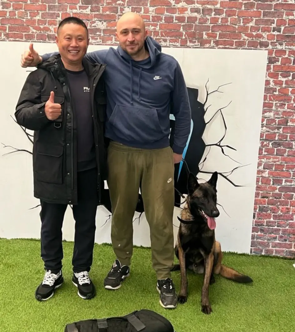 Two men standing with thumbs up next to a seated black and brown dog on artificial grass against a brick and cracked wall backdrop.