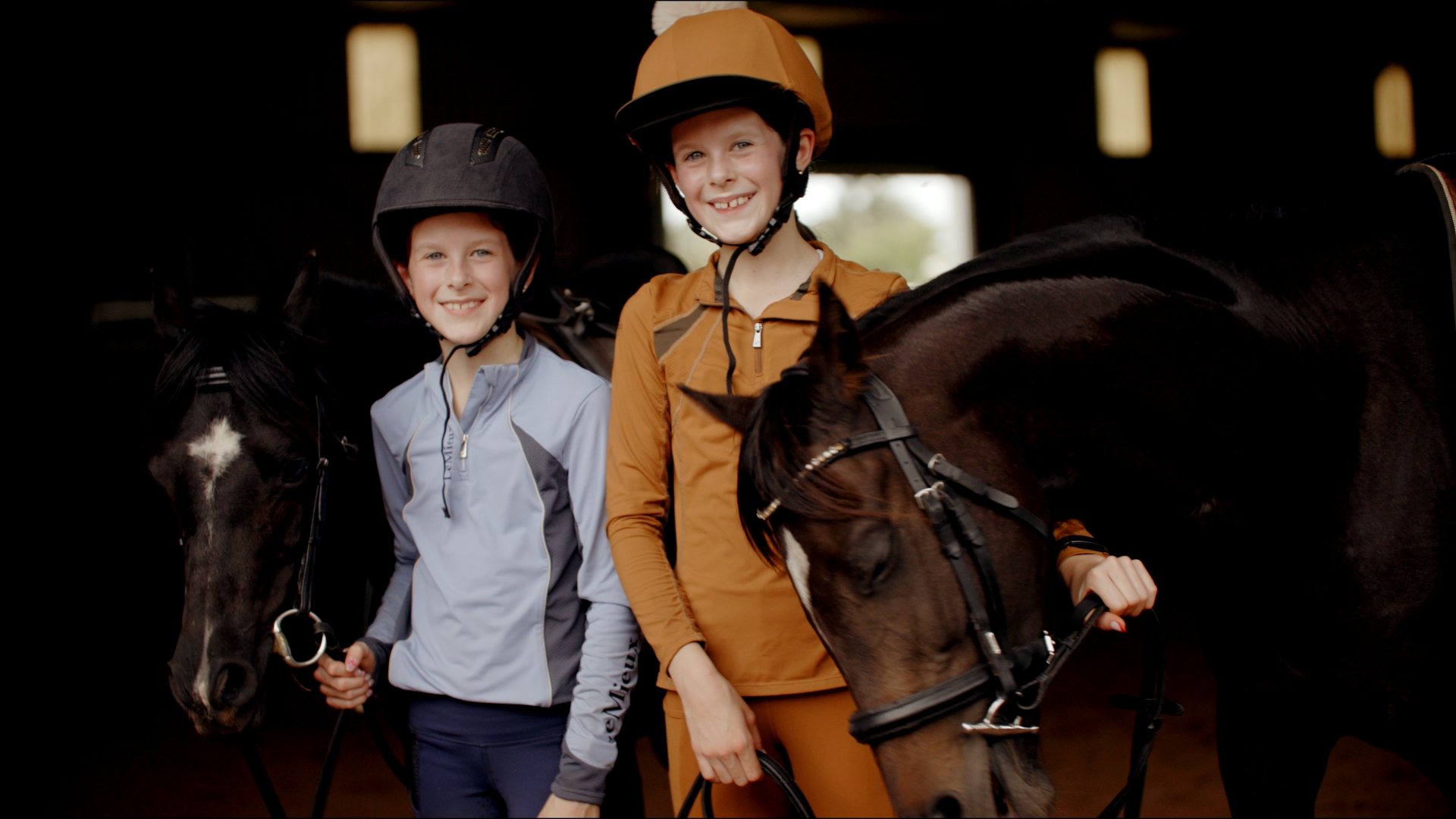 Two children wearing riding helmets standing with their horses inside a stable.